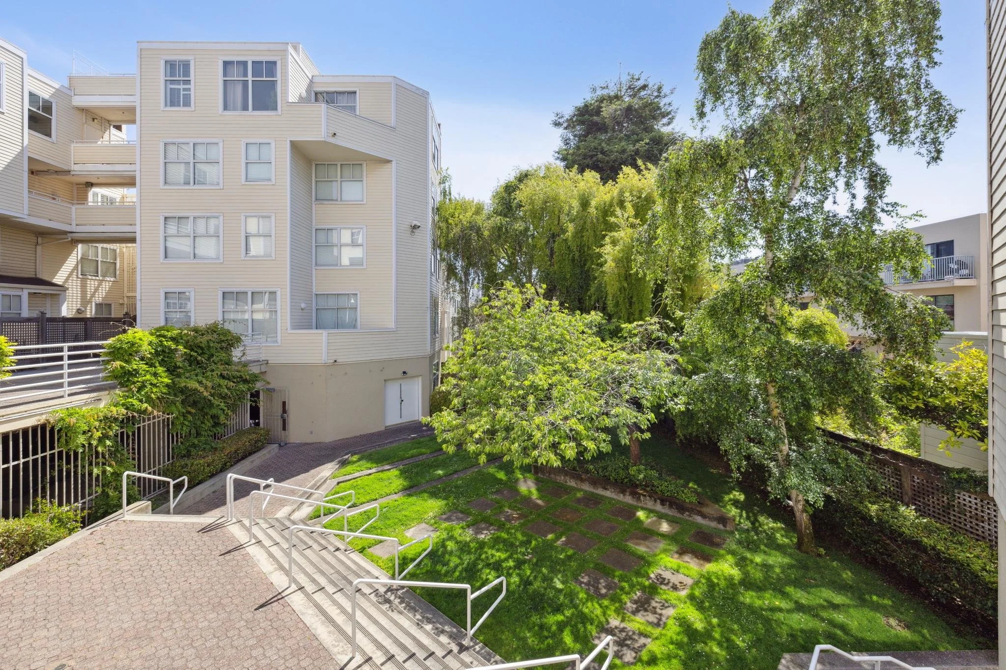 A residential courtyard with a winding pathway, greenery, and multi-story apartment buildings on a sunny day.