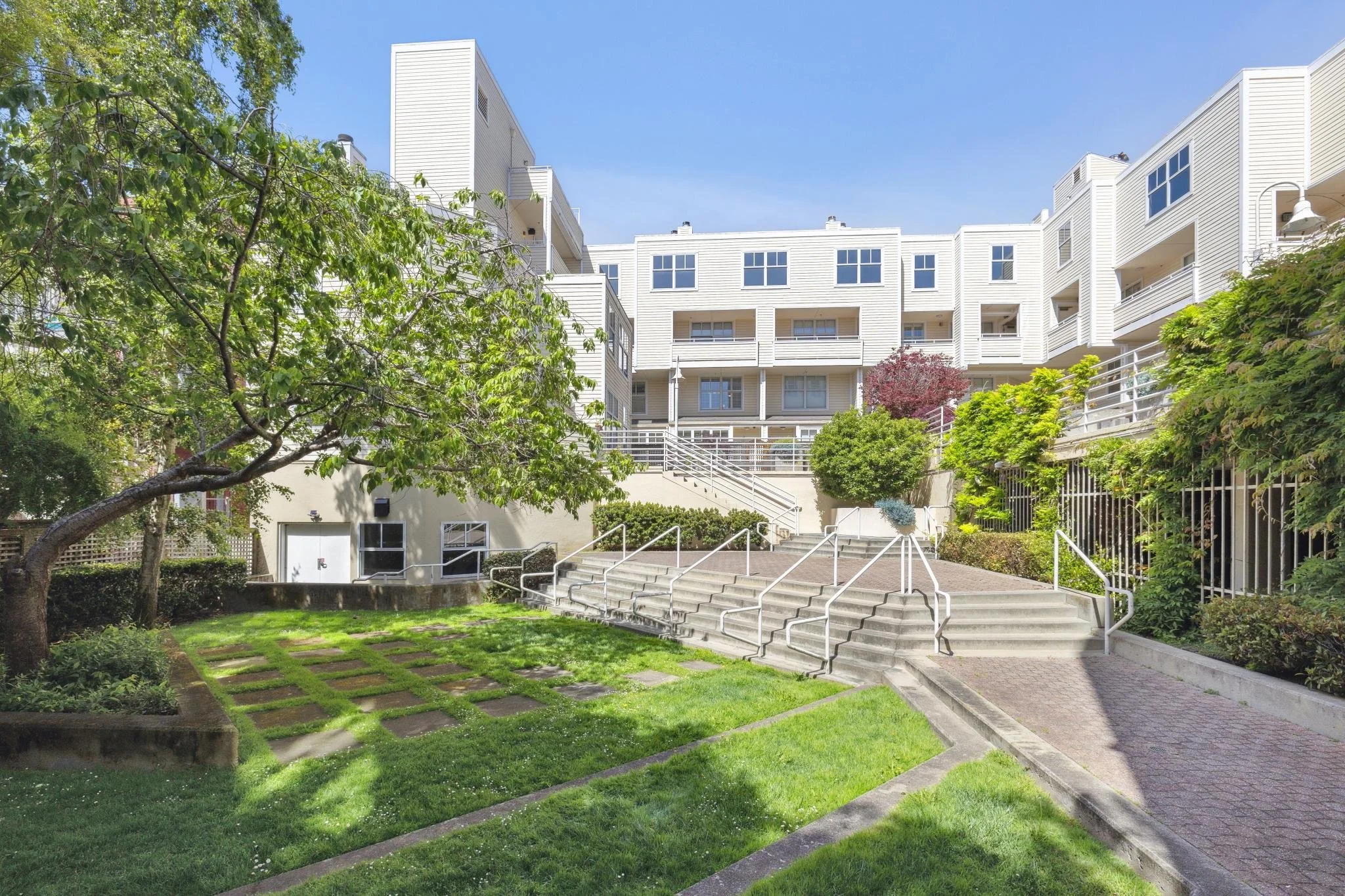 A modern multi-story apartment building with a white exterior, surrounded by a well-maintained garden with stairs and walkways, green trees, and shrubs, under a clear blue sky.