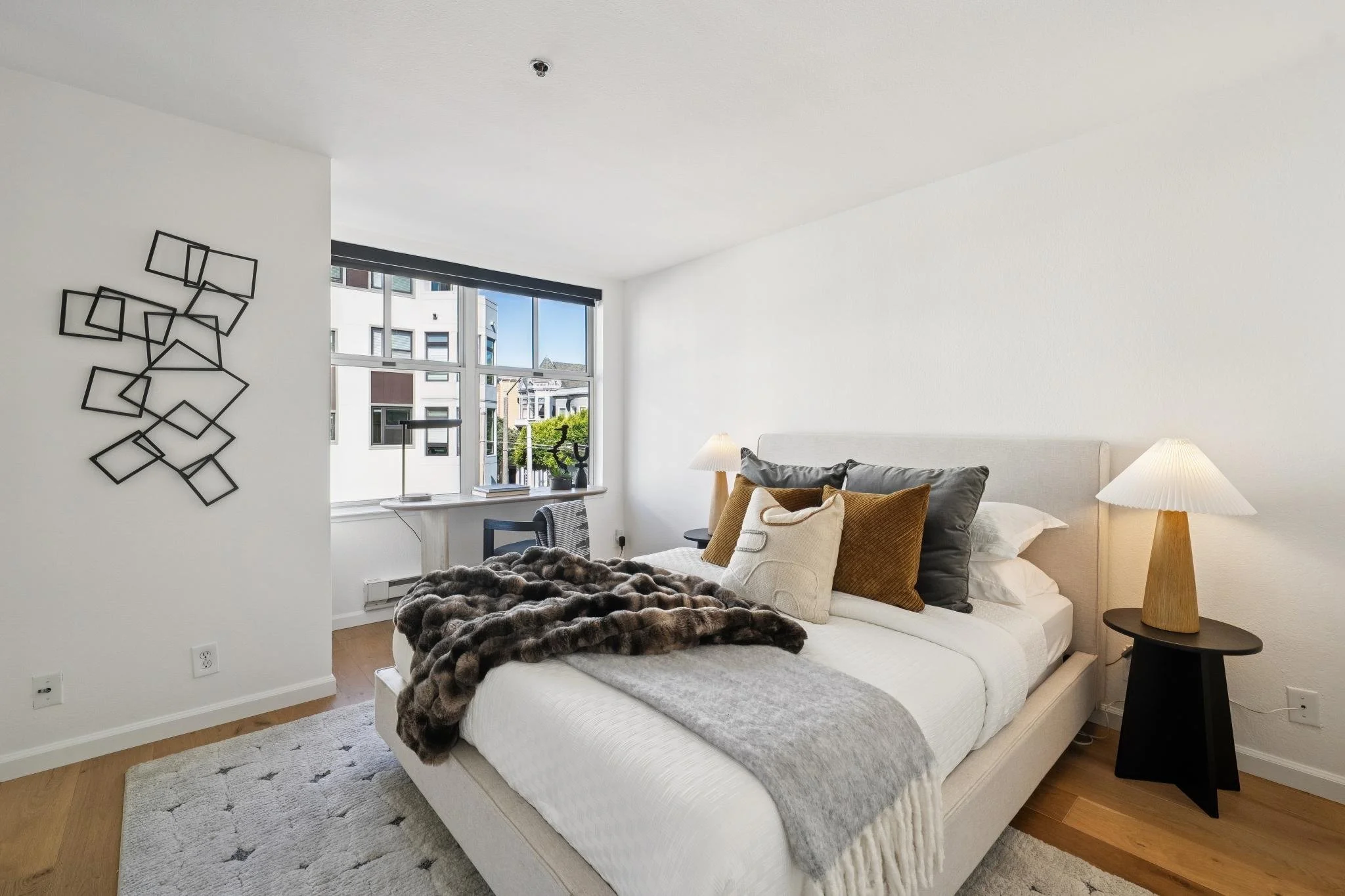 Modern bedroom with large window, white walls, beige bed with gray and brown pillows, dark fur throw, black side table, white lampshades, and abstract black metal wall art.