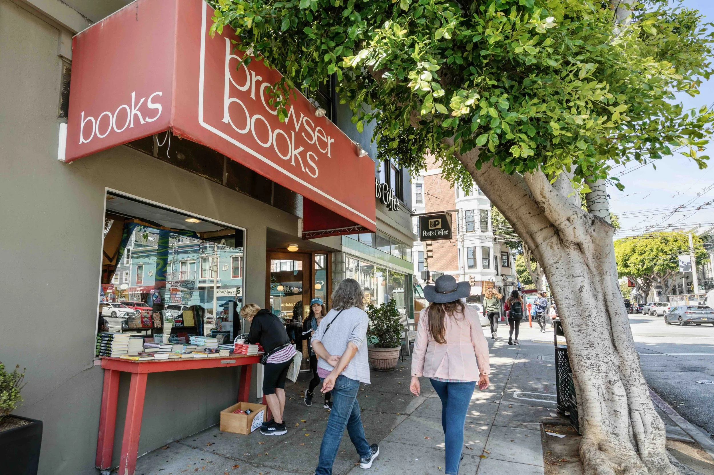 People walking along a city sidewalk past a bookstore with a red canopy, displaying books outside, under a large leafy tree on a sunny day.