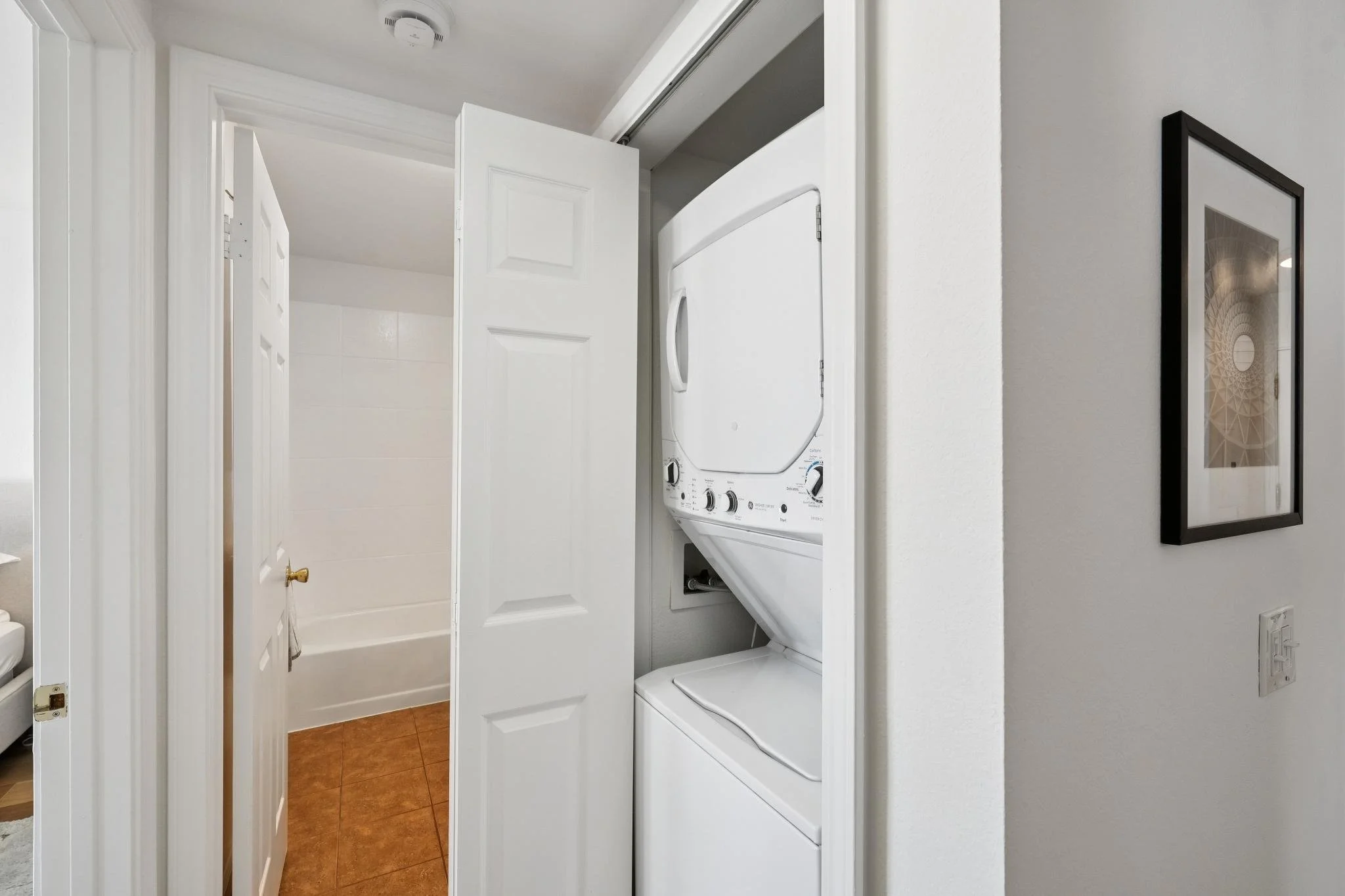 A laundry closet with a stacked washer and dryer behind a partially open door, with a framed picture on the wall to the right.