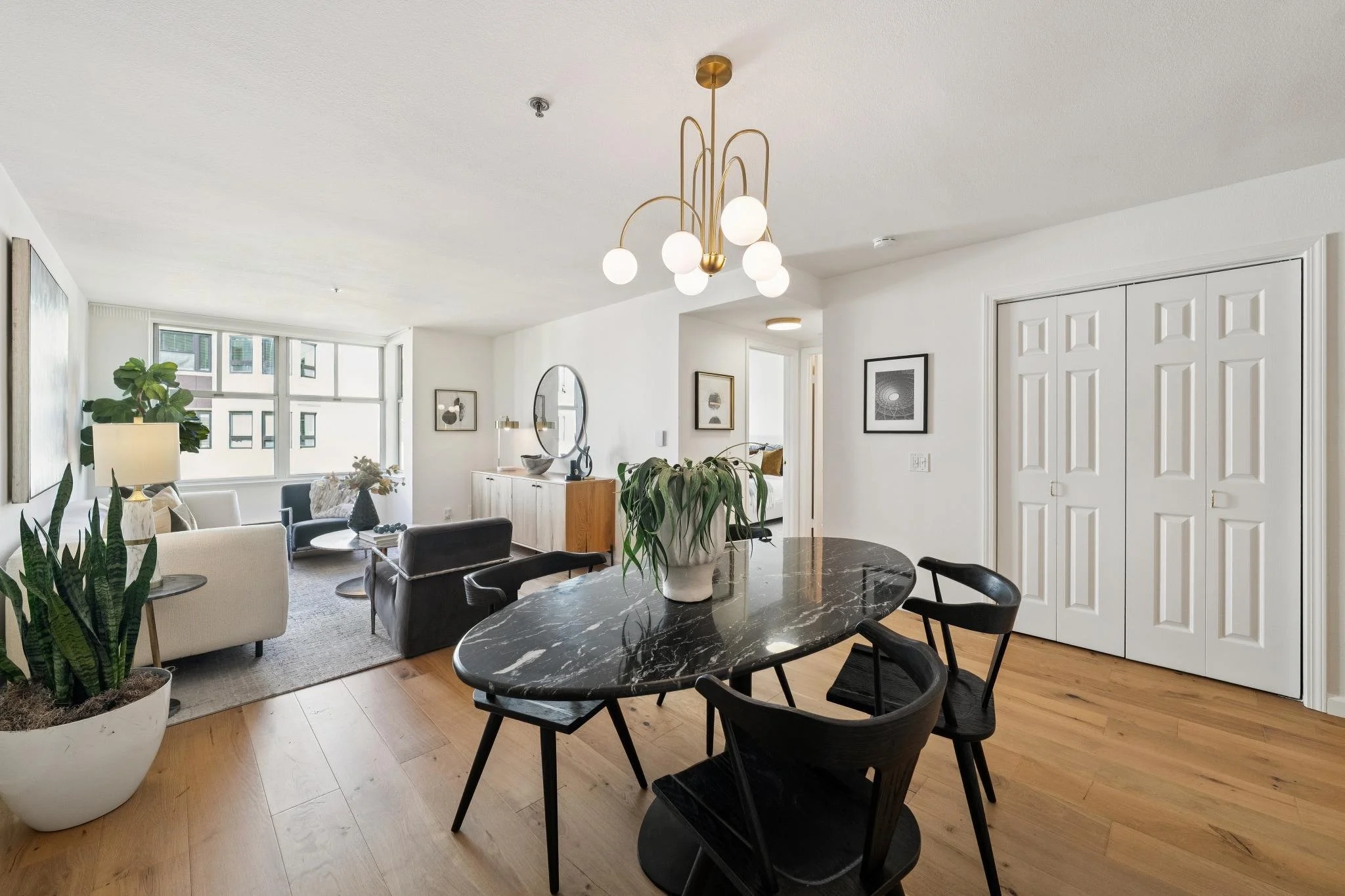 Open living and dining area with modern decor, large windows, potted plants, a chandelier, and framed artwork.
