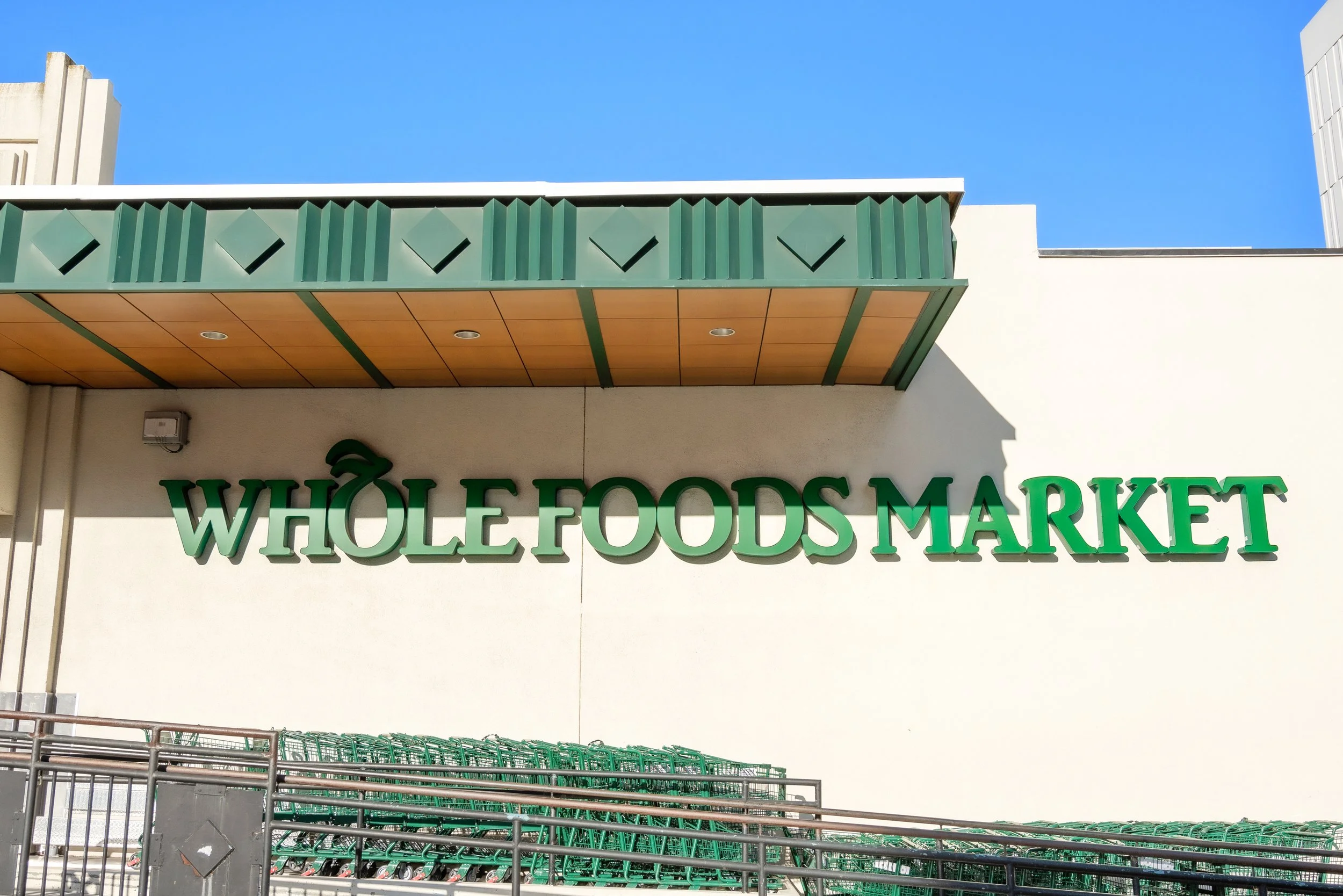 Exterior of a Whole Foods Market store with green lettering and a modern architectural design, shopping carts in the foreground, bright blue sky in the background.