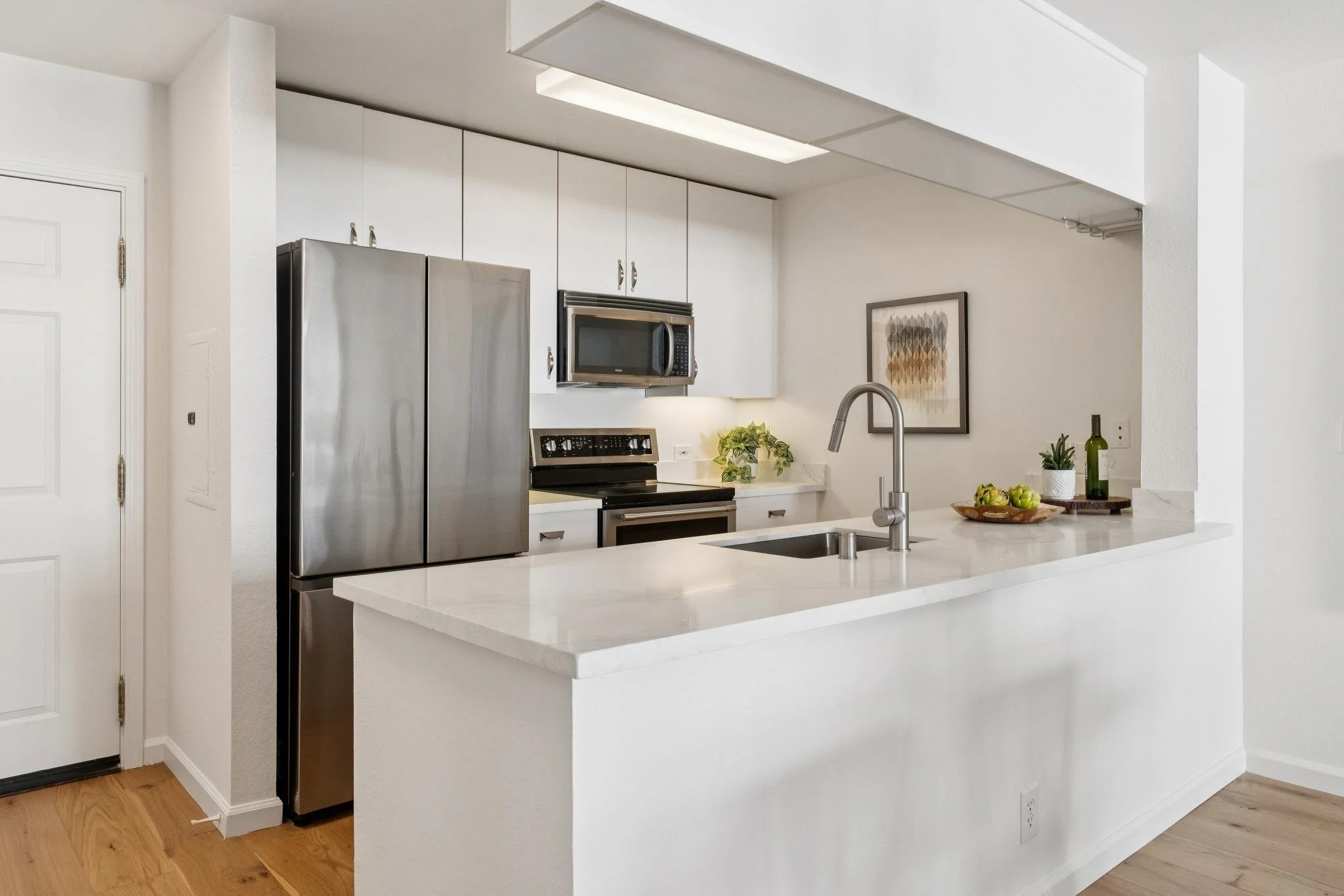 A modern kitchen with white cabinets, stainless steel refrigerator and microwave, white countertop with plants, wood flooring, and artwork on the wall.
