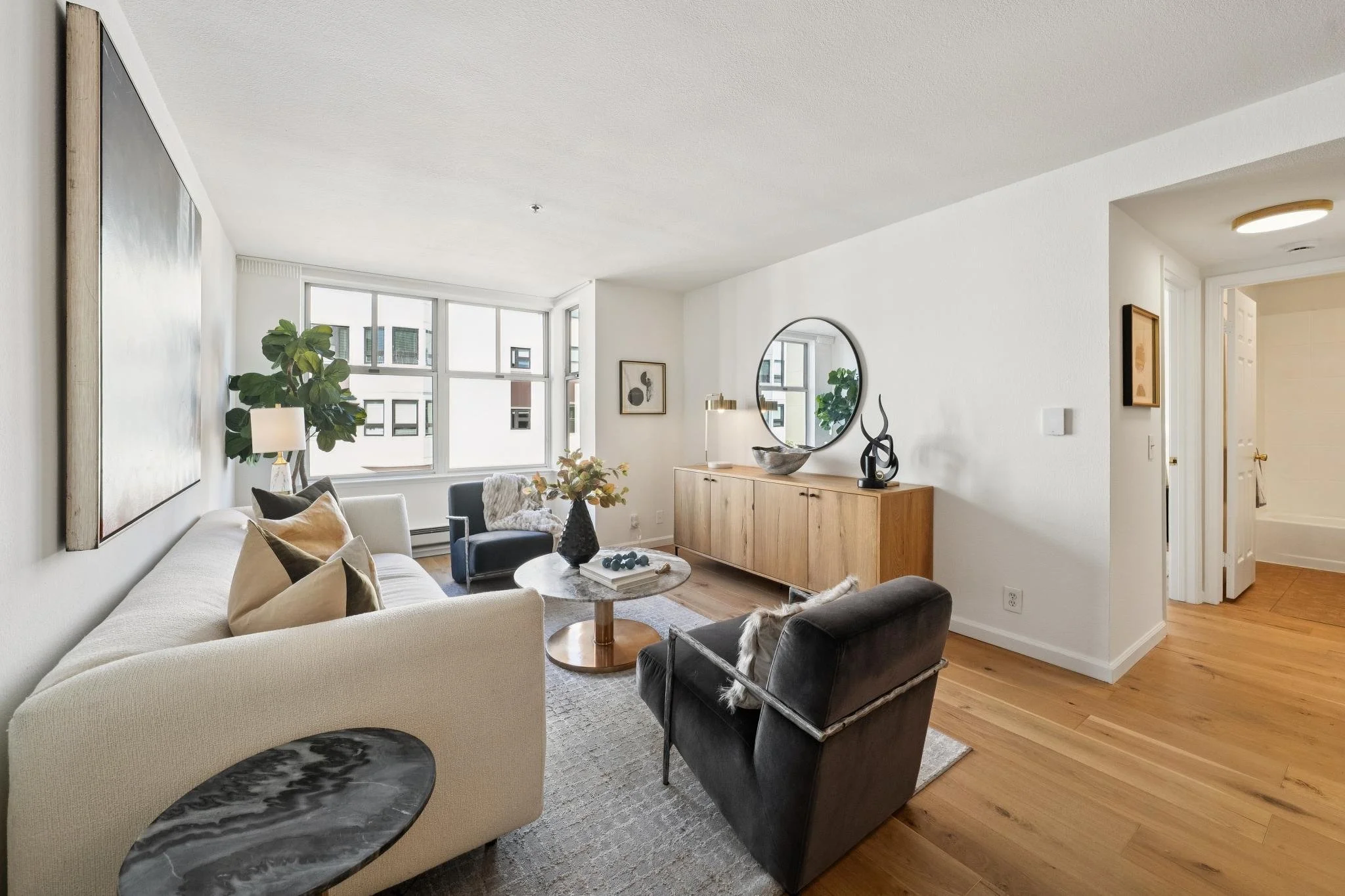 Living room with a large window, white walls, a cream sofa with pillows, black and beige chairs, a wooden sideboard with decorative items, a mirror, and a potted plant.