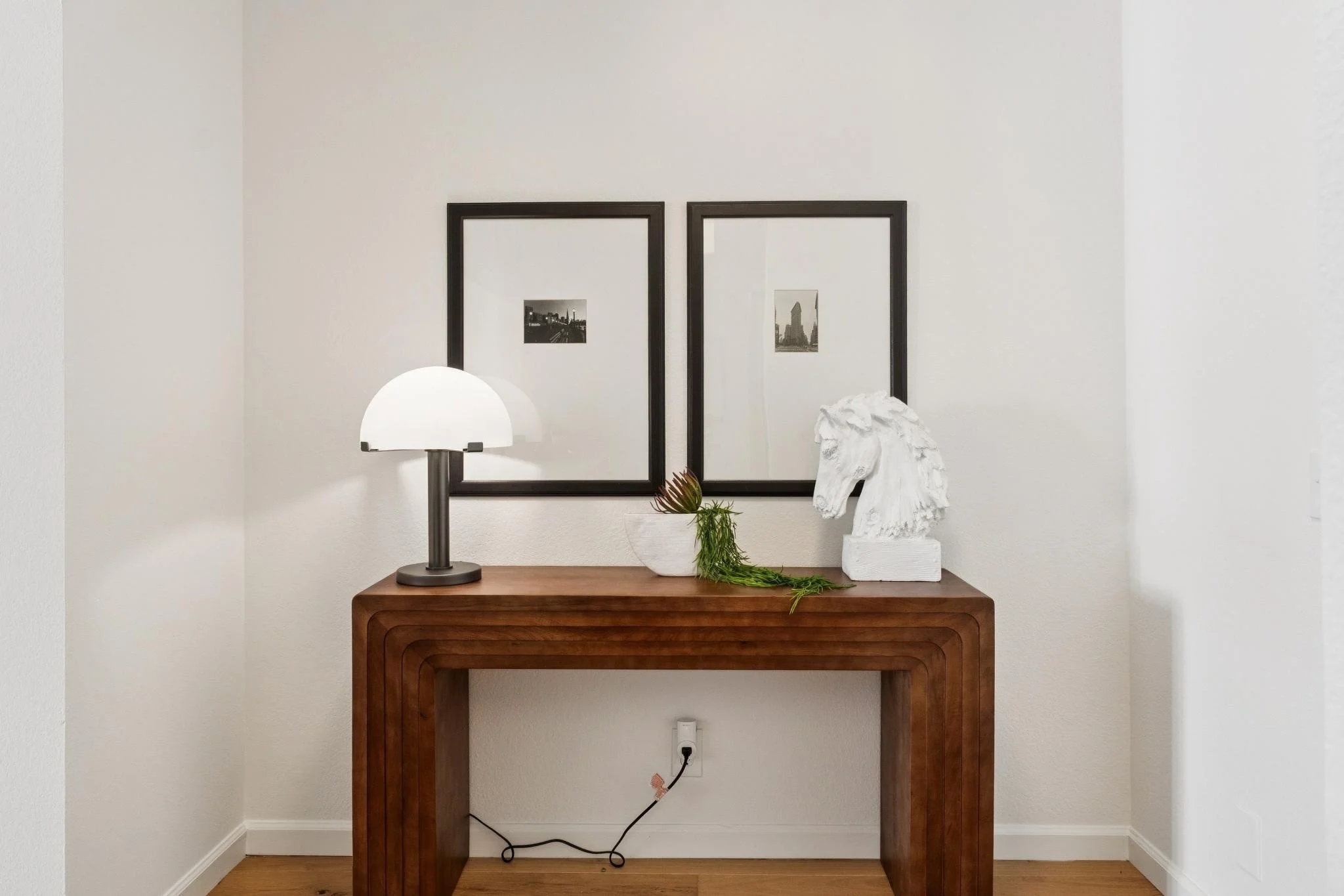 A wooden console table with decorative items including a white lamp, a white sculpture of a horse's head, and a plant in a white pot. Behind the table, there are two framed black-and-white cityscape photographs on a white wall.