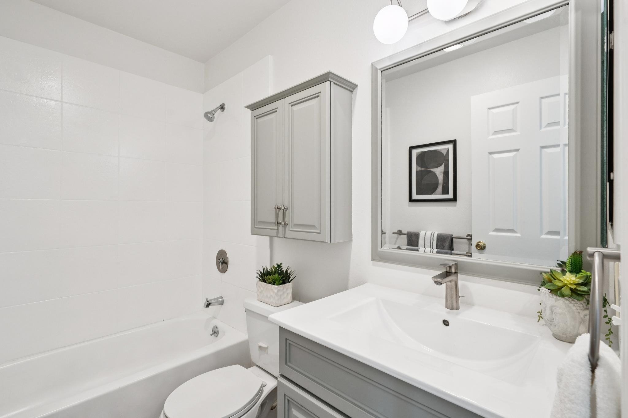 A modern bathroom with white walls and a large mirror, gray cabinetry, potted plants, a bathtub, and a towel rack.