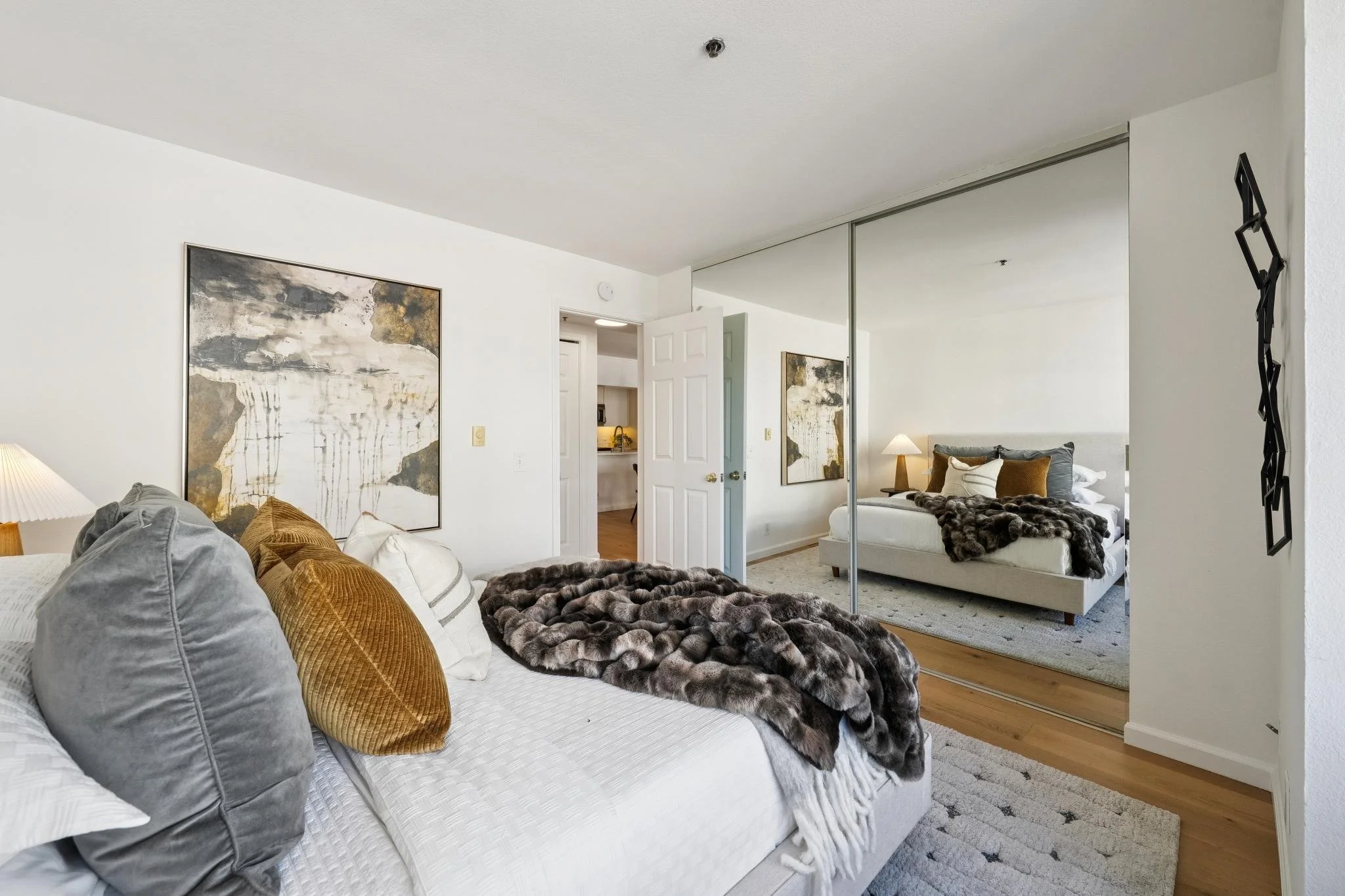 A modern bedroom with a daytime view, featuring a white bed with gray, gold, and white pillows, a plush fur throw, artwork on the walls, a mirror closet door reflecting the room, and a doorway leading to a small kitchen area.