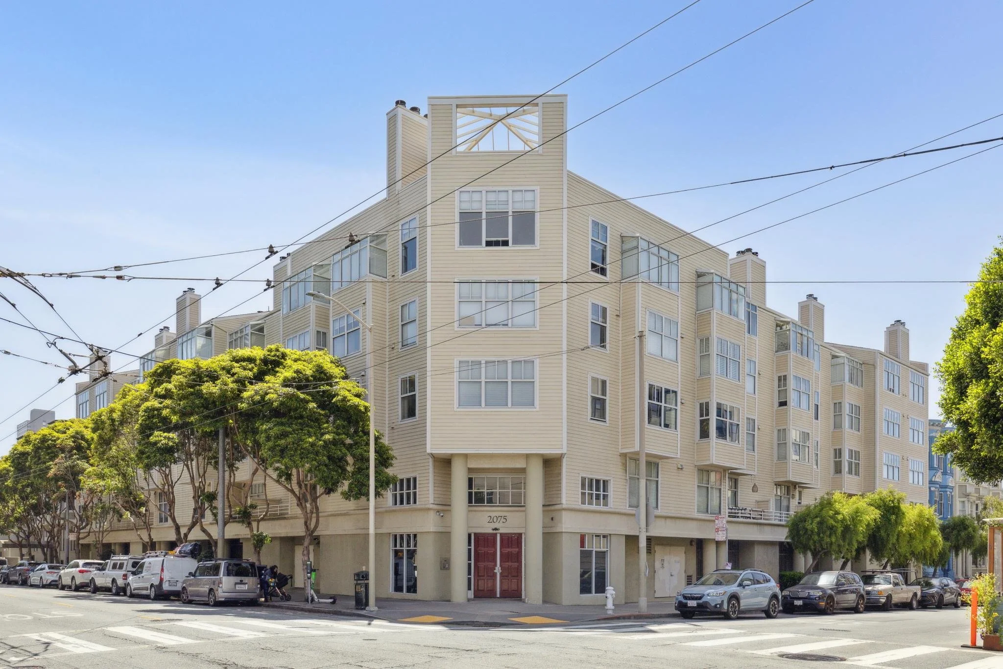 A multi-story beige residential building with numerous windows, located on a city street with parked cars and lined with green trees. Overhead utility lines are visible in the sky.