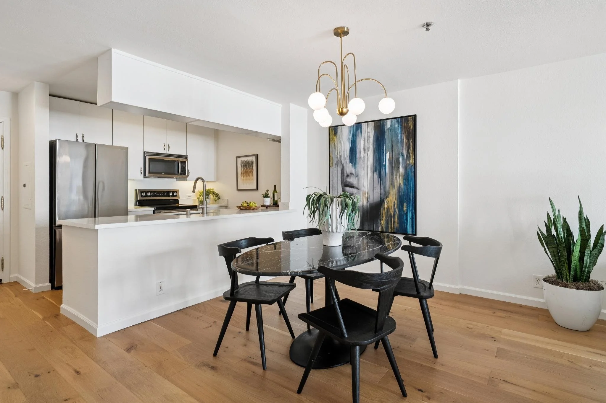 Modern dining area with black table and chairs, large indoor plant, white wall, large abstract artwork, and modern chandelier with round globe lights.
