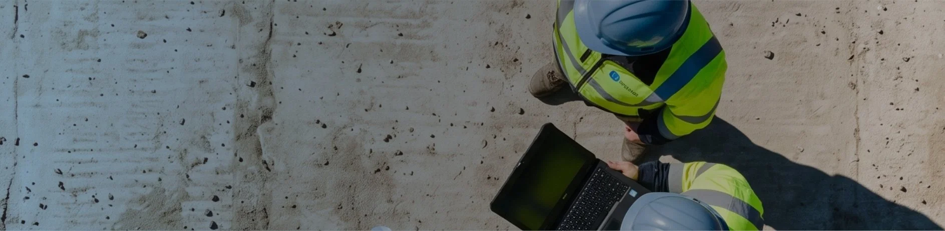 Two construction workers wearing safety helmets and high-visibility jackets working on a concrete surface with a laptop.