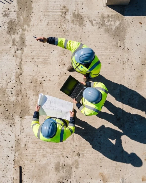 Three construction workers wearing yellow reflective safety vests and blue hard hats standing on a construction site, examining blueprints and a laptop.