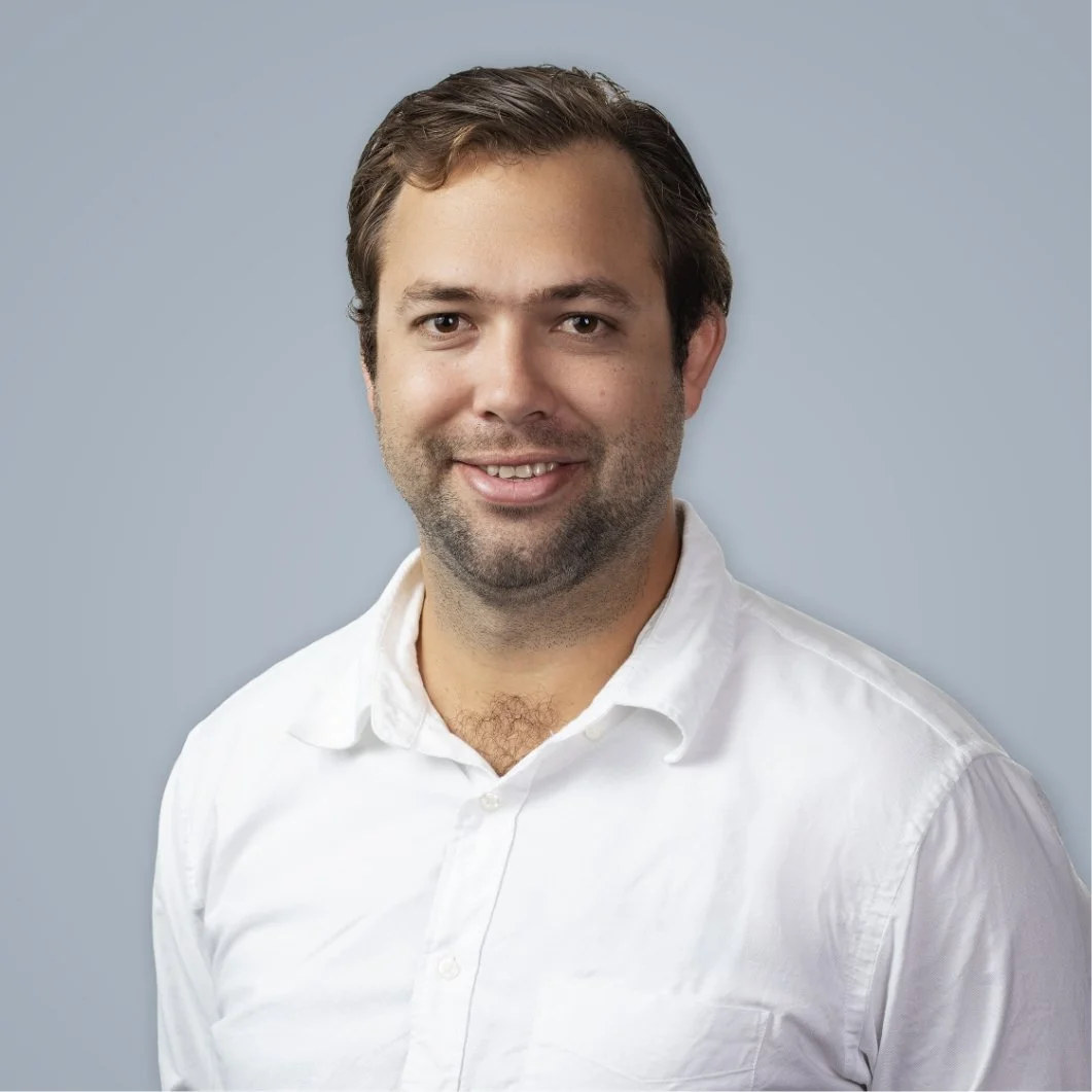 Portrait of a man with short, dark hair and a beard, wearing a white button-up shirt, smiling against a light gray background.