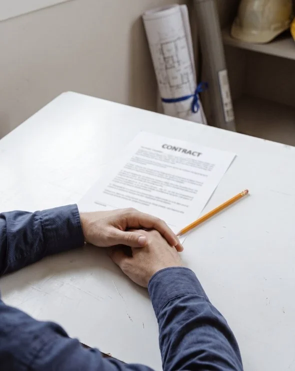 Hands folded on a white table next to a contract and a yellow pencil in an office or workspace setting.