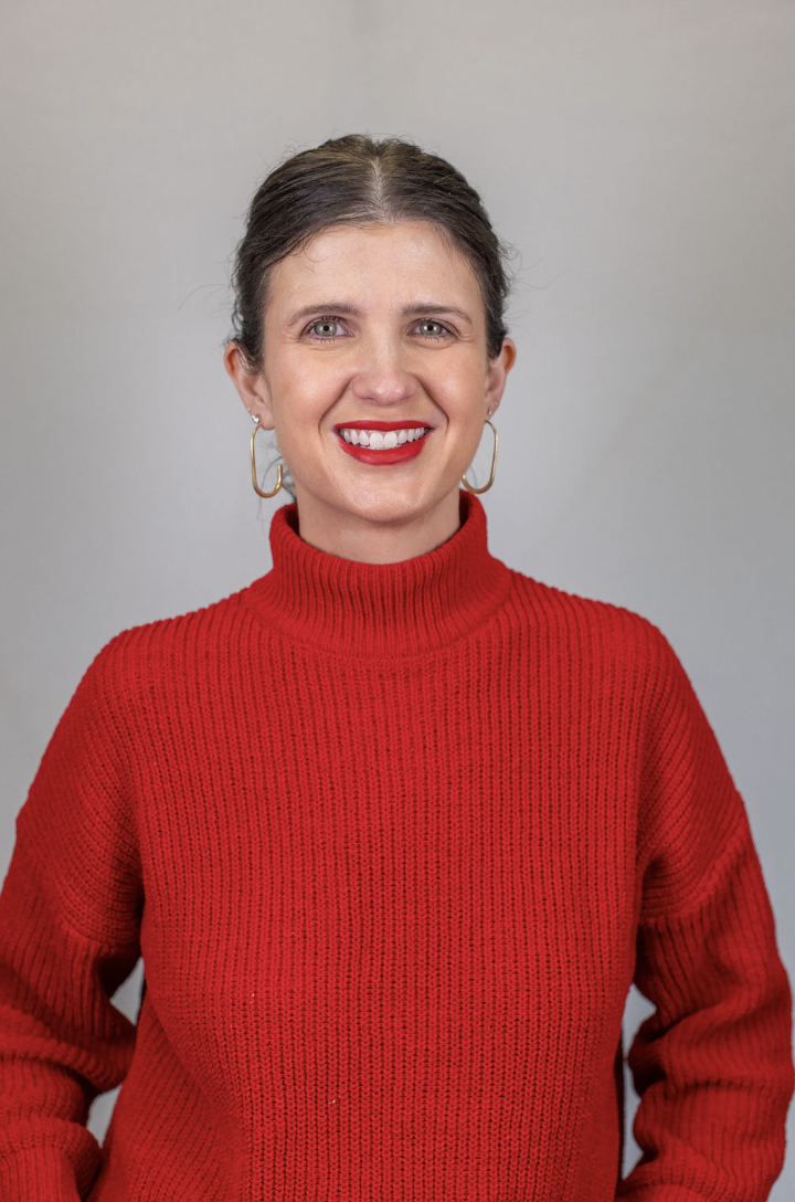 A woman with brown hair pulled back, wearing a red turtleneck sweater and gold hoop earrings, smiling at the camera against a plain gray background.