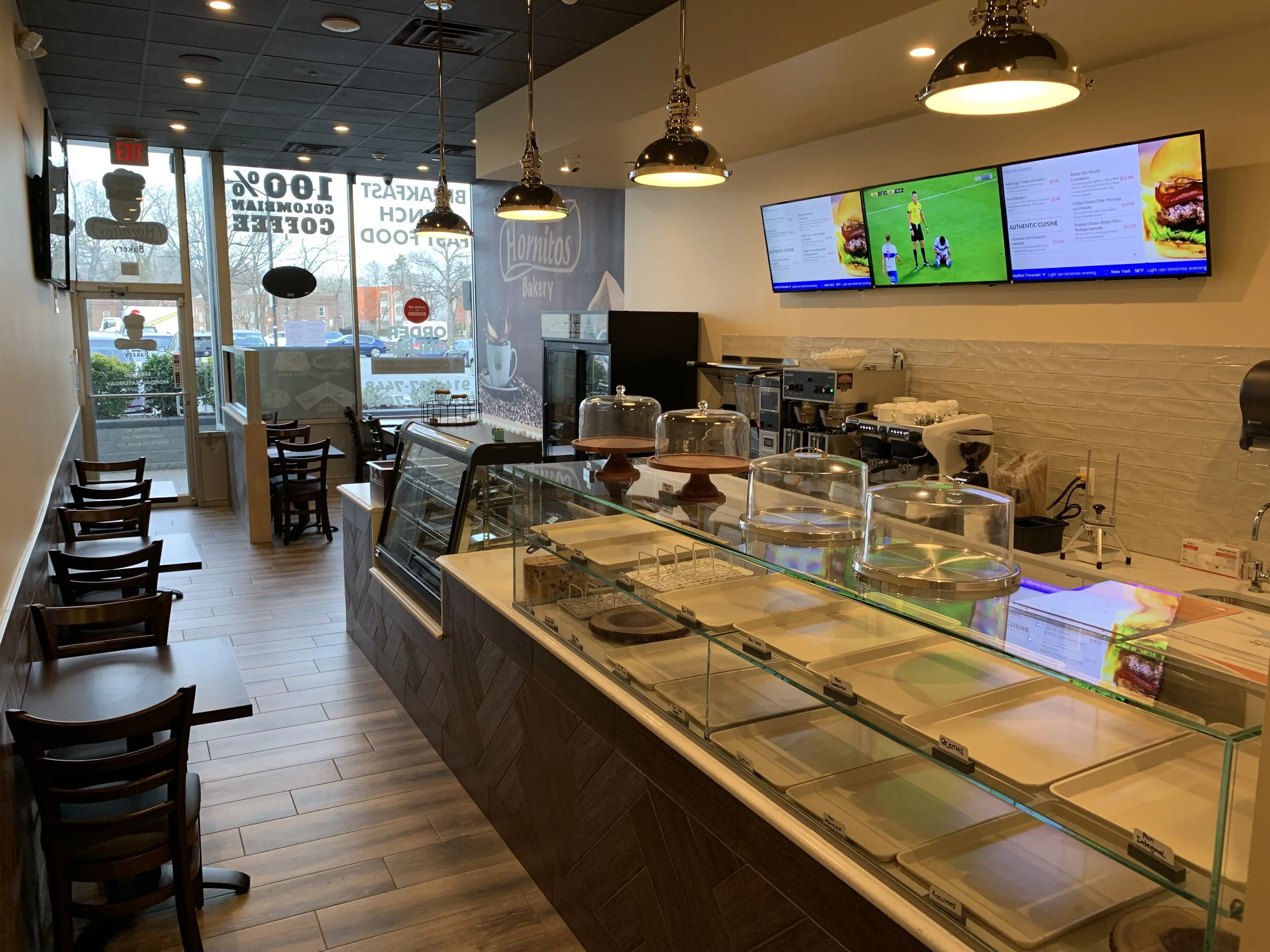 Interior of a bakery or cafe with a glass display counter filled with empty trays, a menu on digital screens showing food options and prices, and seating with dark chairs. There are three black pendant lights hanging from the ceiling and the front large window shows the outside parking lot and trees.