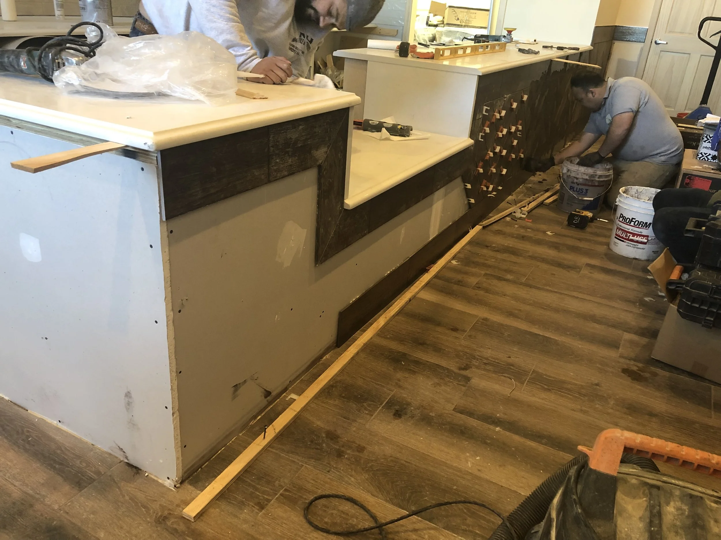 Two workers are installing or repairing a kitchen counter. One is bending down working near the cabinet, and the other is standing at the far end of the counter. There are tools and construction materials scattered around.