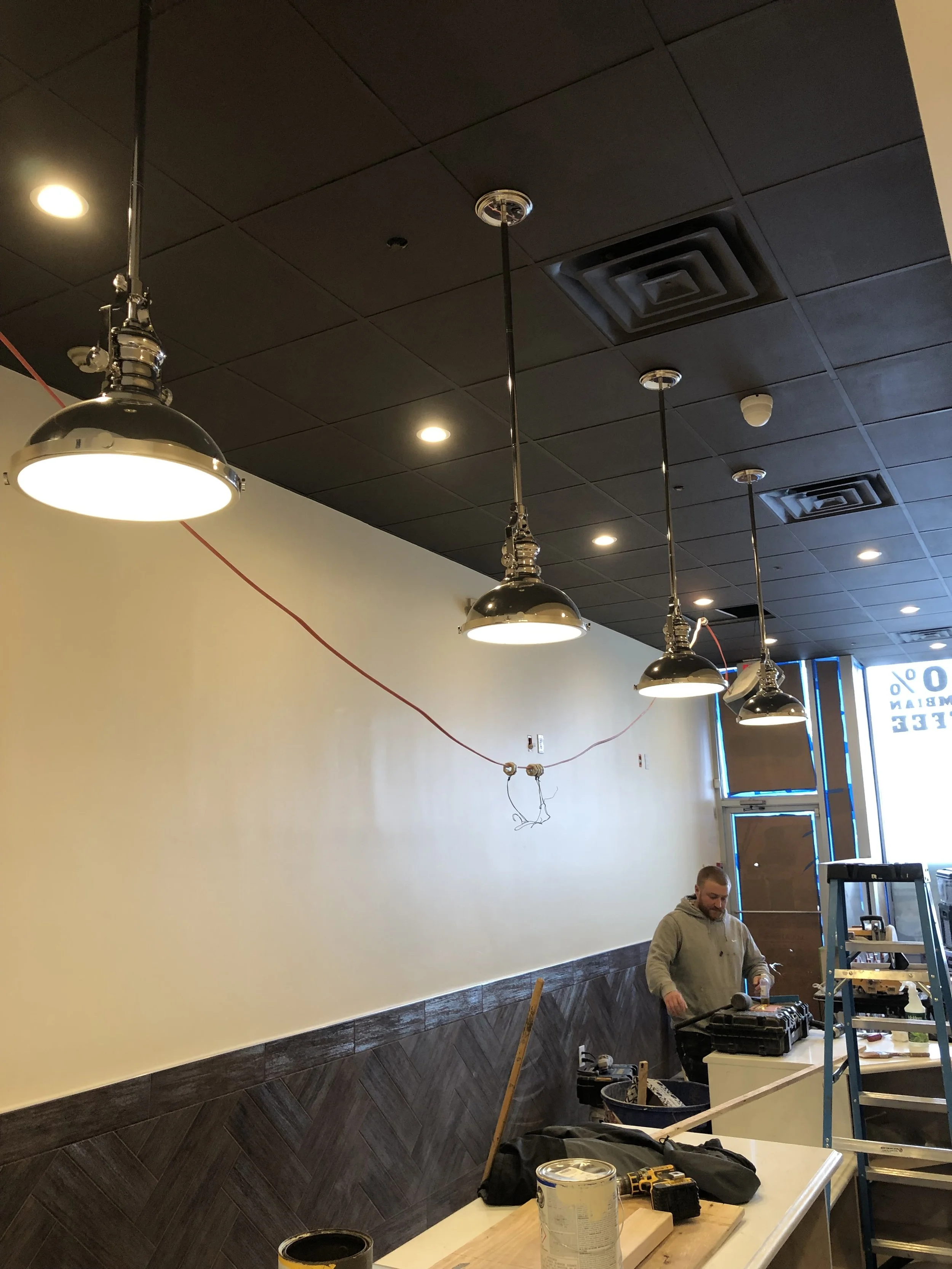 Interior of a restaurant under renovation with black pendant lights hanging from a black ceiling, a man working at a table with tools and materials, and a wall with a beige upper section and dark wood lower section, with a light fixture and small smoke detector on the ceiling.