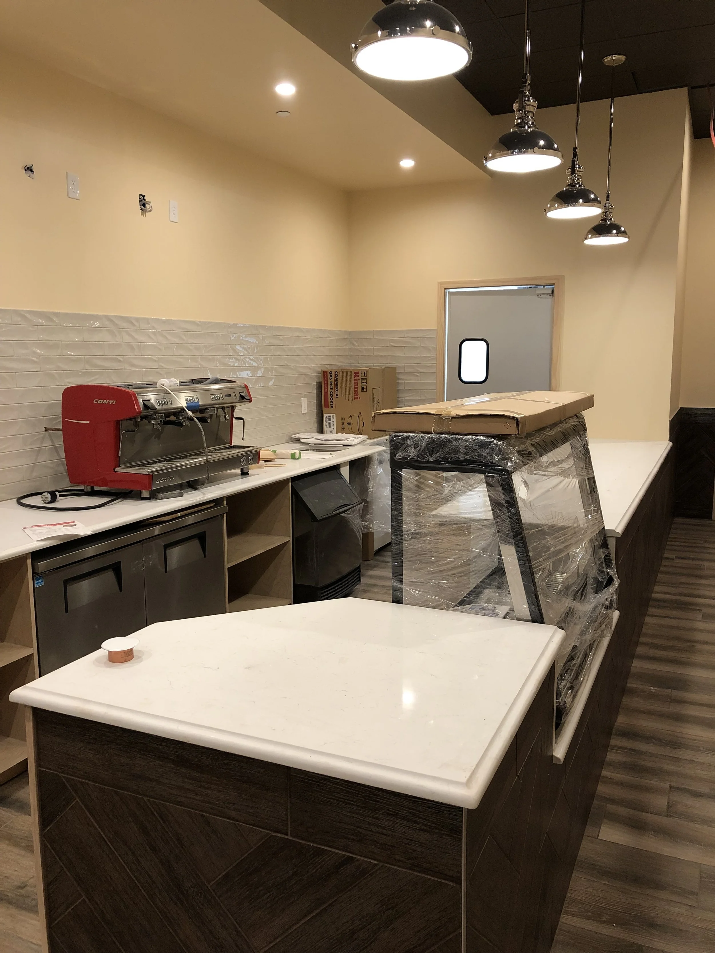 An unfinished food service or coffee counter with a red espresso machine, boxes, and other supplies, surrounded by tan walls and light fixtures.