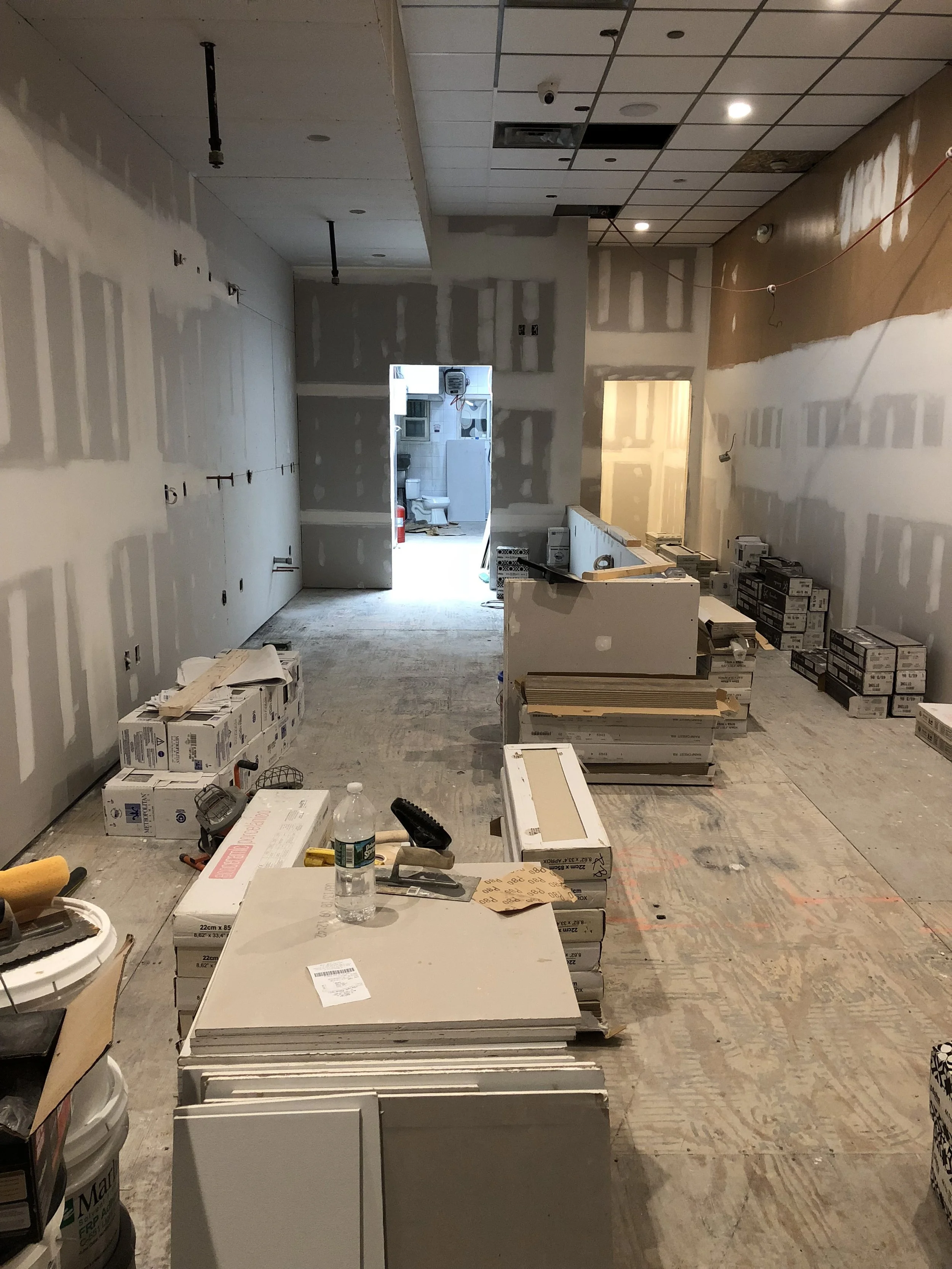 A room under construction or renovation with drywall and ceiling panels, construction materials, boxes, and tools scattered around.