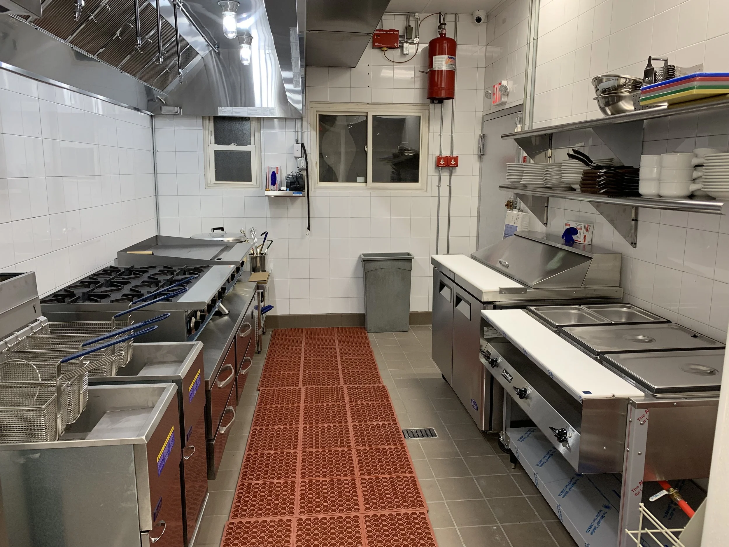 Commercial kitchen with stainless steel appliances, shelves with dishes, fire extinguisher, and a red rubber mat on the floor.