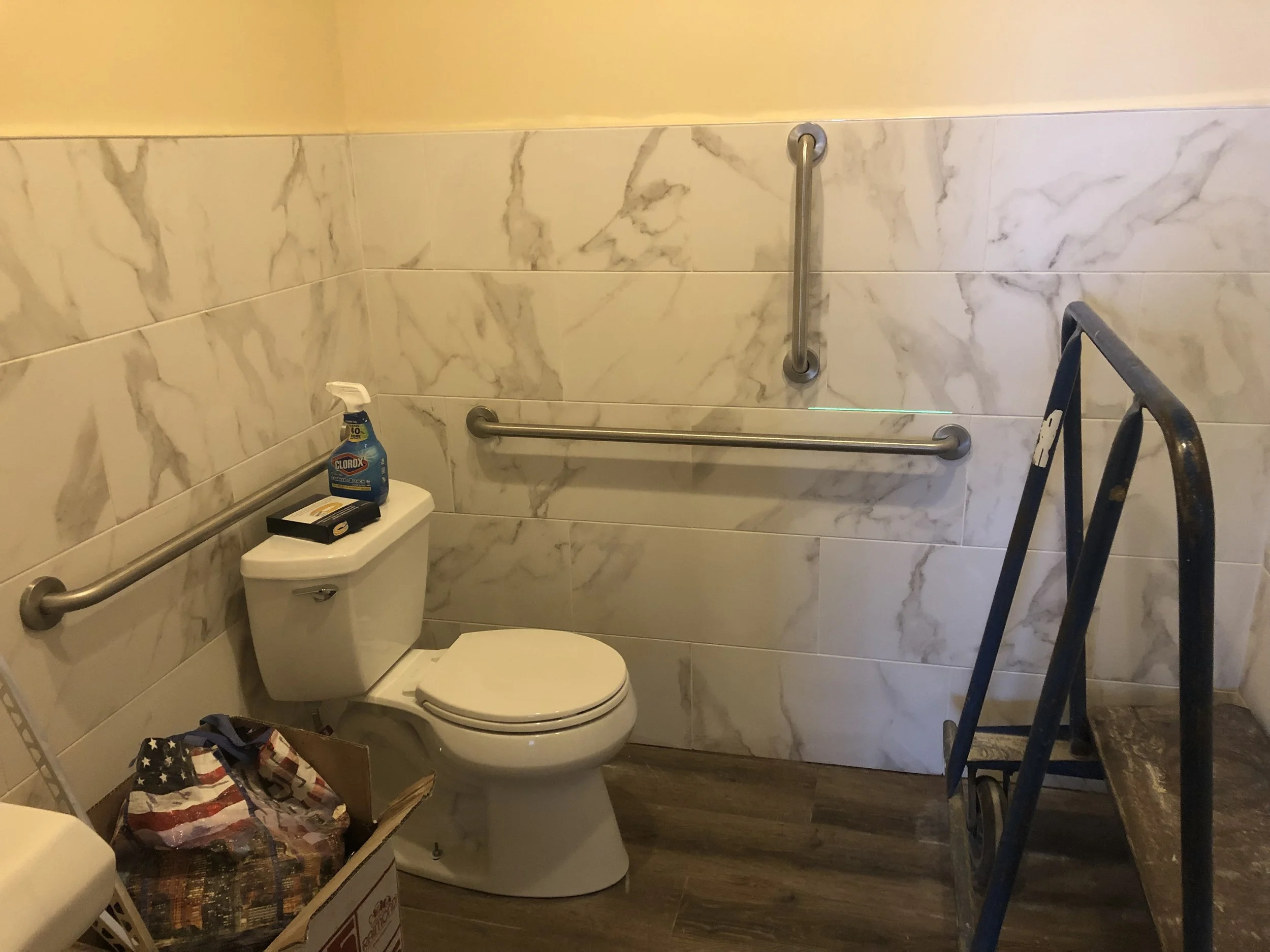 A bathroom with marble wall tiles, a toilet, a bottle of Clorox disinfectant spray, a cardboard box filled with American flag-printed fabric, and a blue step ladder.