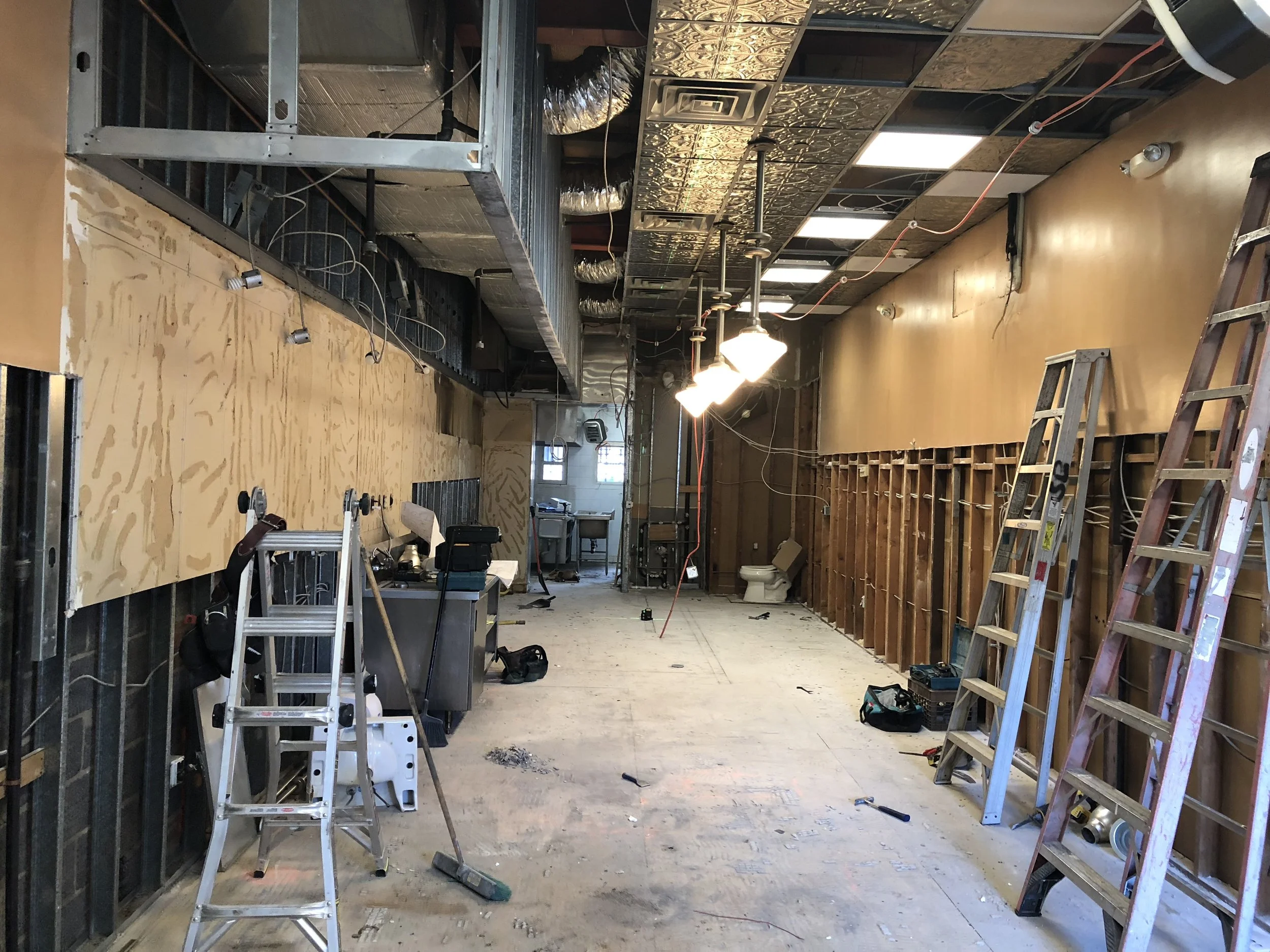 Interior of a construction or renovation site with ladders, tools, exposed ceiling tiles, ductwork, electrical wires, and unfinished walls.