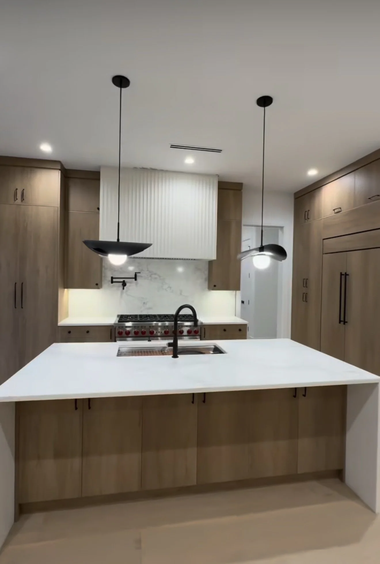Modern kitchen with wooden cabinets, white countertop island, black sink faucet, black pendant lights, white marble backsplash, and stainless steel stove.