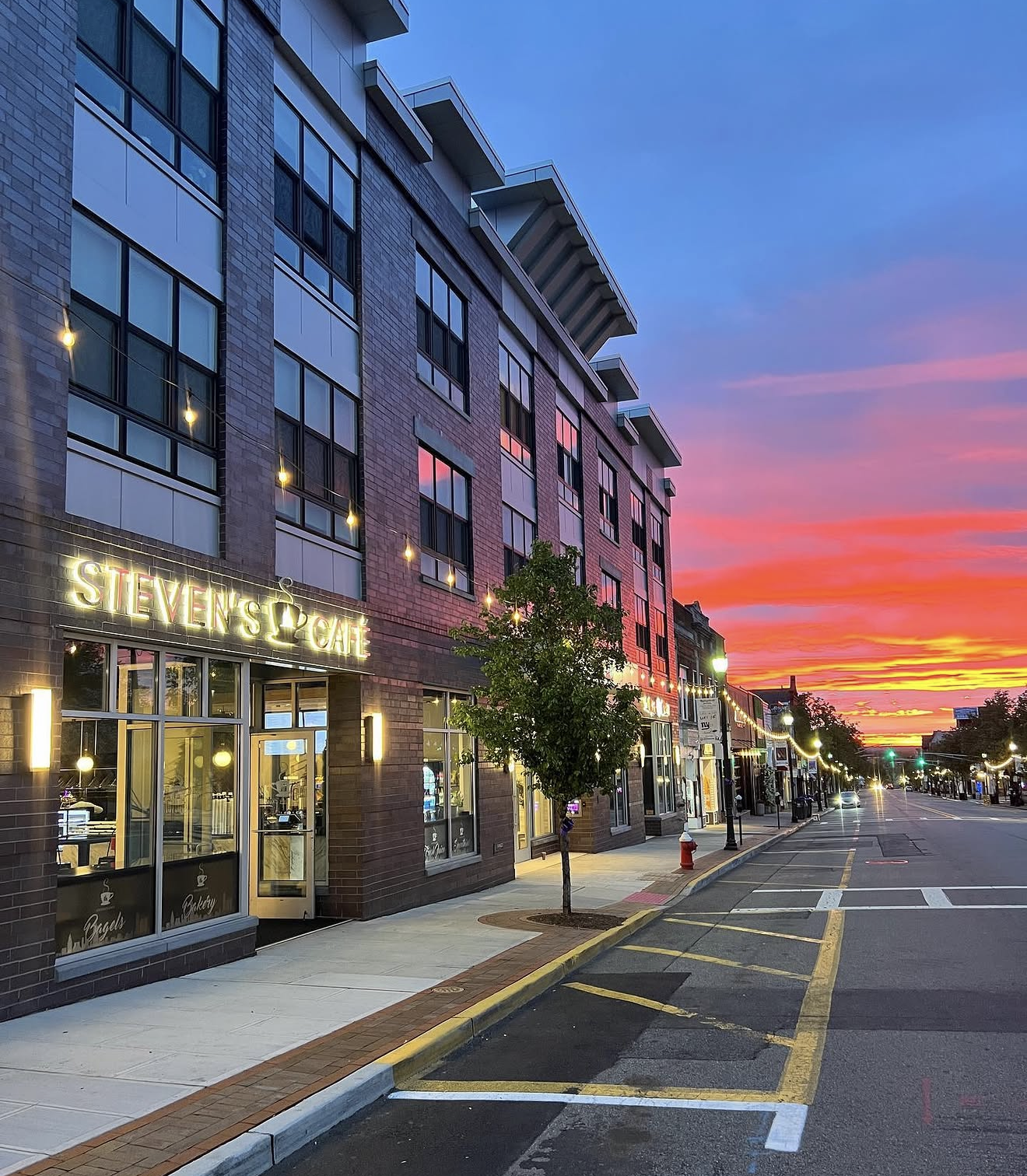 Empty street with shops and café, colorful sunset sky with pink, purple, orange, and yellow clouds, building with neon signs, trees, streetlights, and cars parked along the curb.