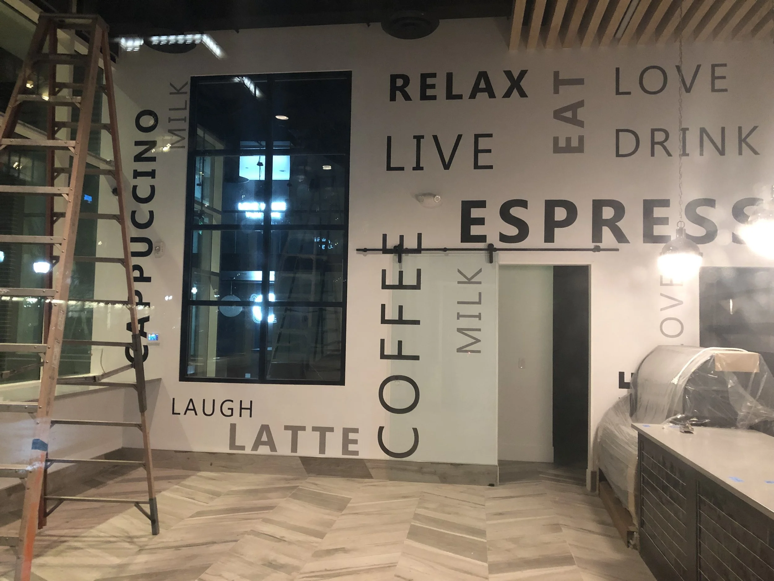 Interior of a cafe or coffee shop with large black text on the wall displaying words like 'relax,' 'love,' 'live,' 'smile,' and 'eat.' A wooden ladder is positioned on the left side, and a large window or glass door is in the background, with reflections of indoor lighting.