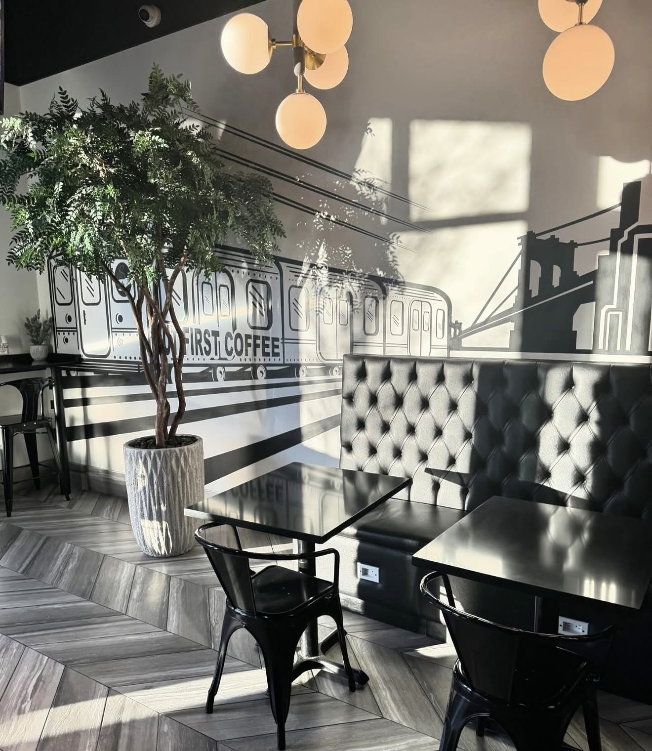 Modern cafe interior with black tables, black chairs, a tufted black booth, a large potted plant, and a mural of a train with the words "First Coffee." The wall features a cityscape with a bridge. There are spherical ceiling lights, and sunlight creates shadows on the wall.