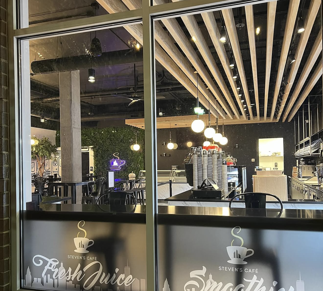 Interior of a modern café seen through a glass window. The café has wooden ceiling slats, pendant lighting, a coffee counter with equipment, and a seating area with tables and chairs. The window has branding for Steven's Café with a coffee cup logo and the words "Fresh Juice" and "Smoothies."