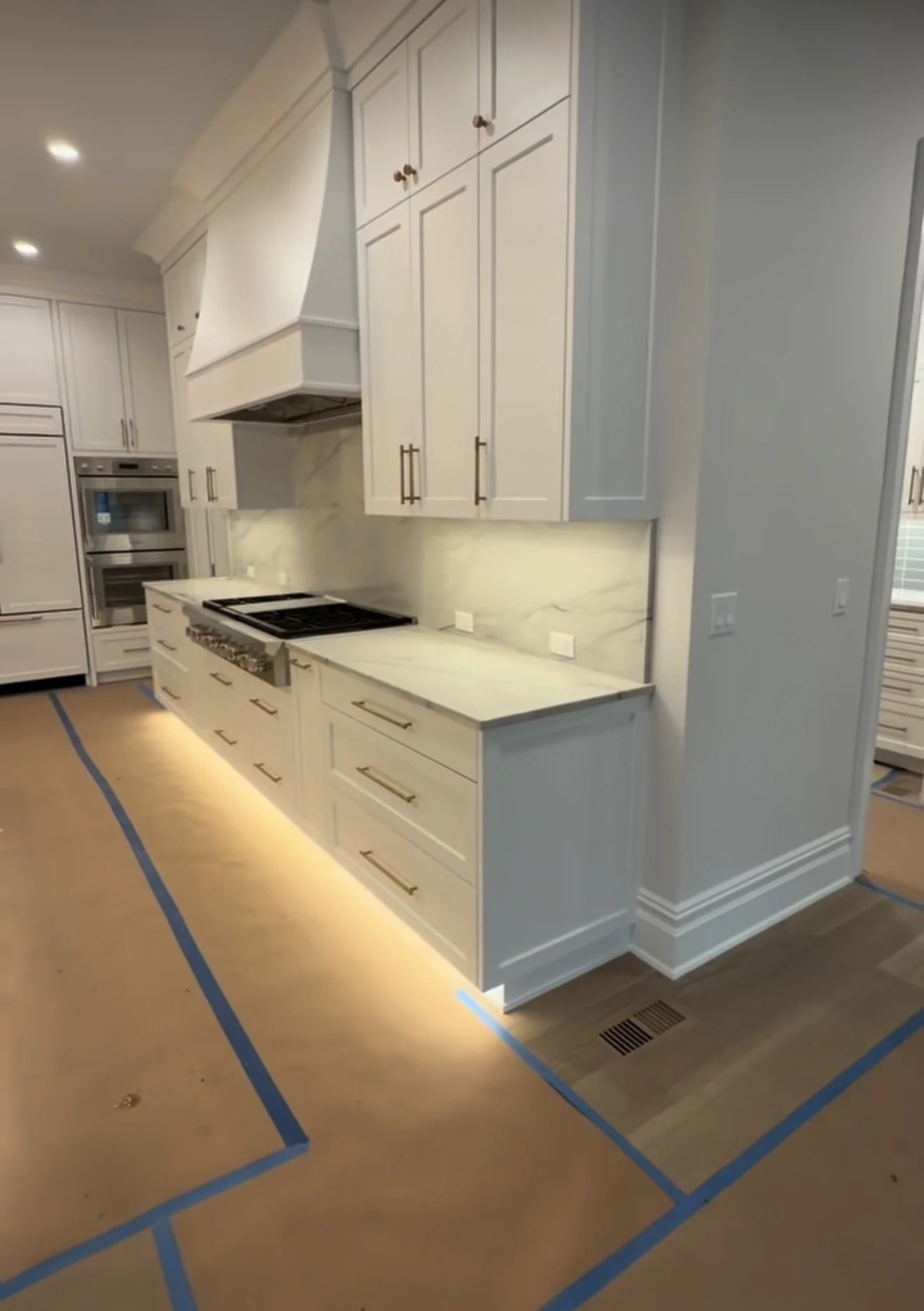 Modern kitchen with white cabinets, marble backsplash, built-in oven, and cooktop, with blue painter's tape on the floor indicating room for flooring installation.