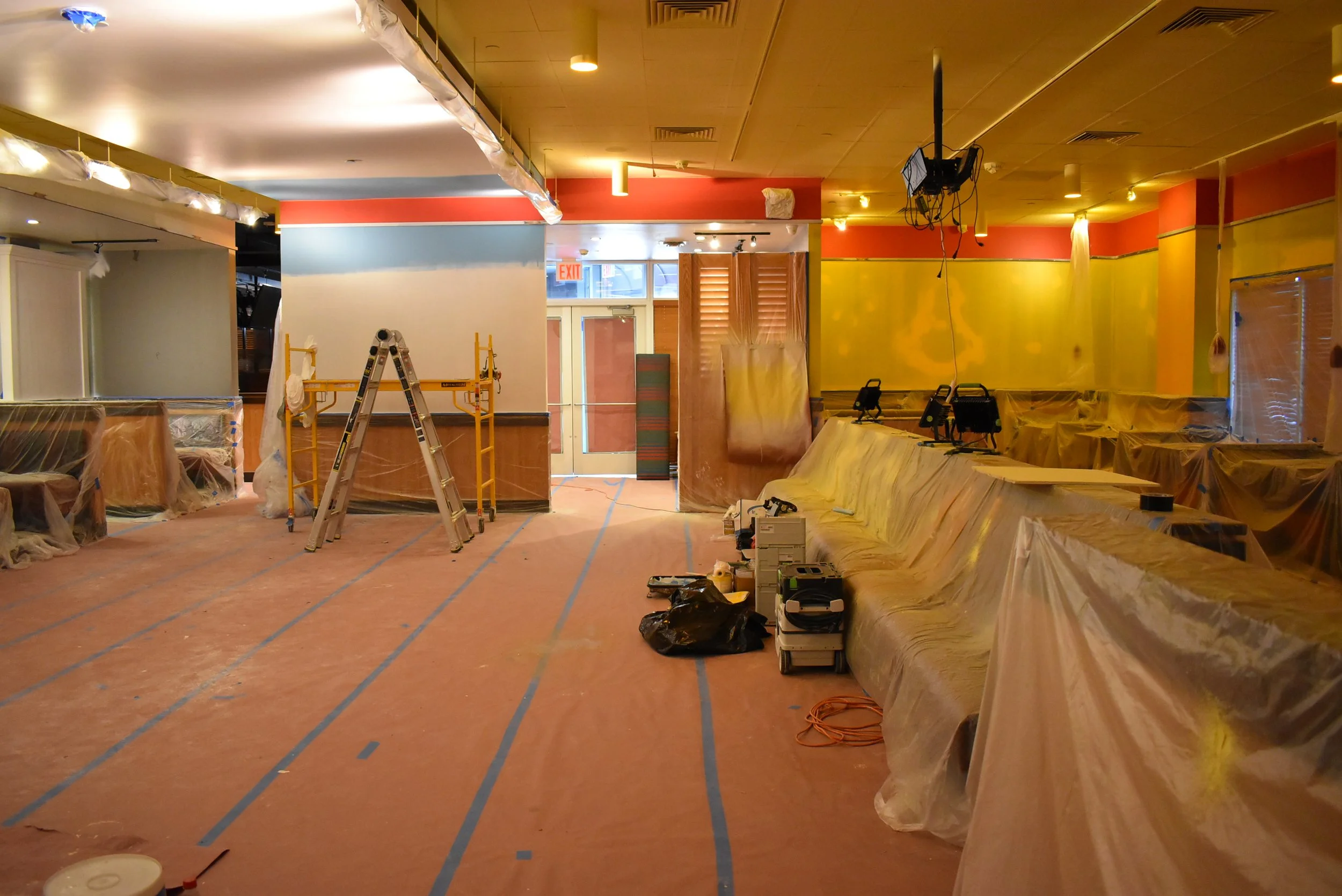 Interior of a restaurant under renovation with plastic-covered furniture, a ladder, tools, and construction materials on the floor.