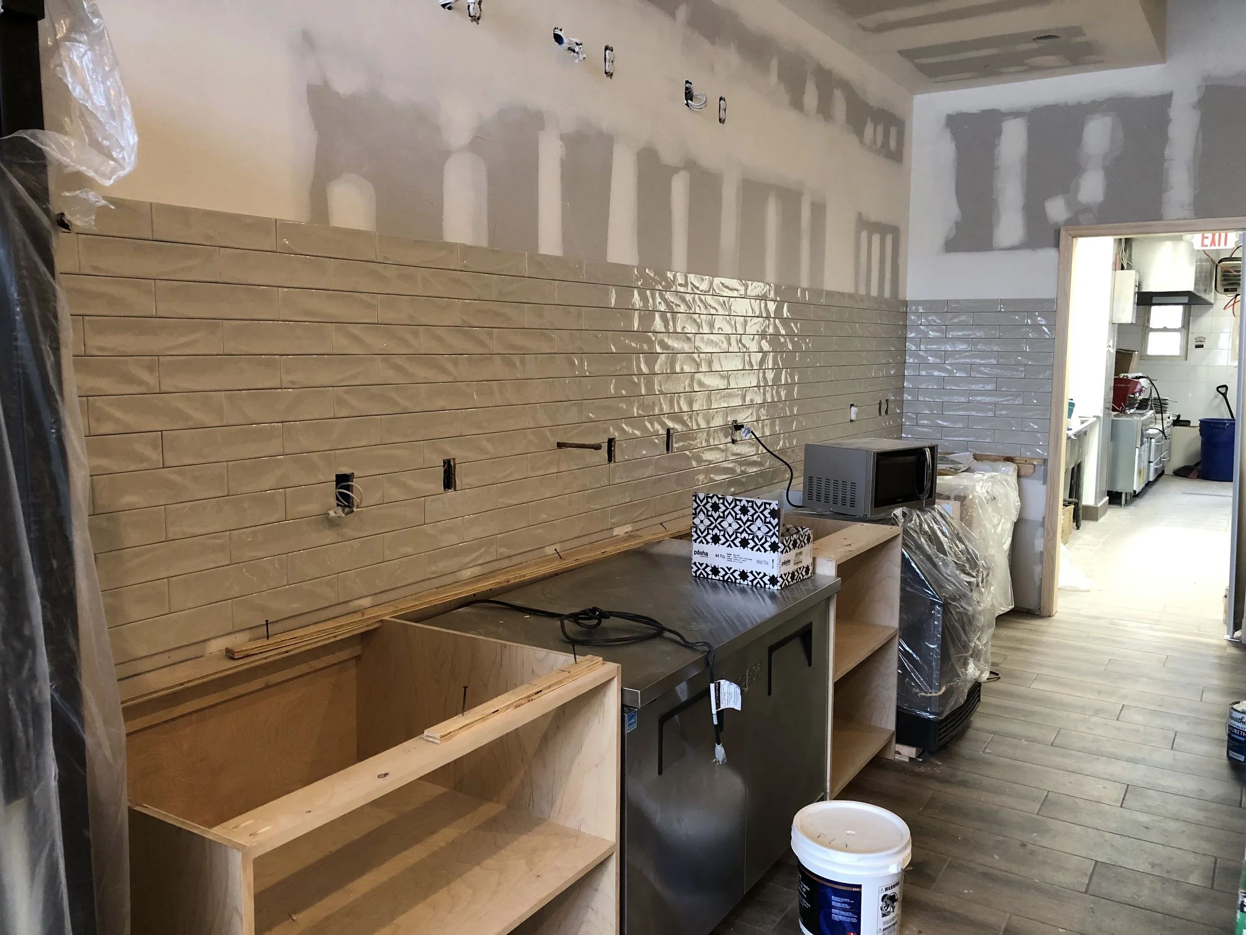 Interior room under renovation with partially tiled wall, electrical outlets, and construction supplies. A back wall is partially tiled with beige glossy tiles, and a wooden shelving unit is present below the tiled wall. Construction tools, a microwave, a bucket, and plastic covering are scattered around, indicating ongoing work.