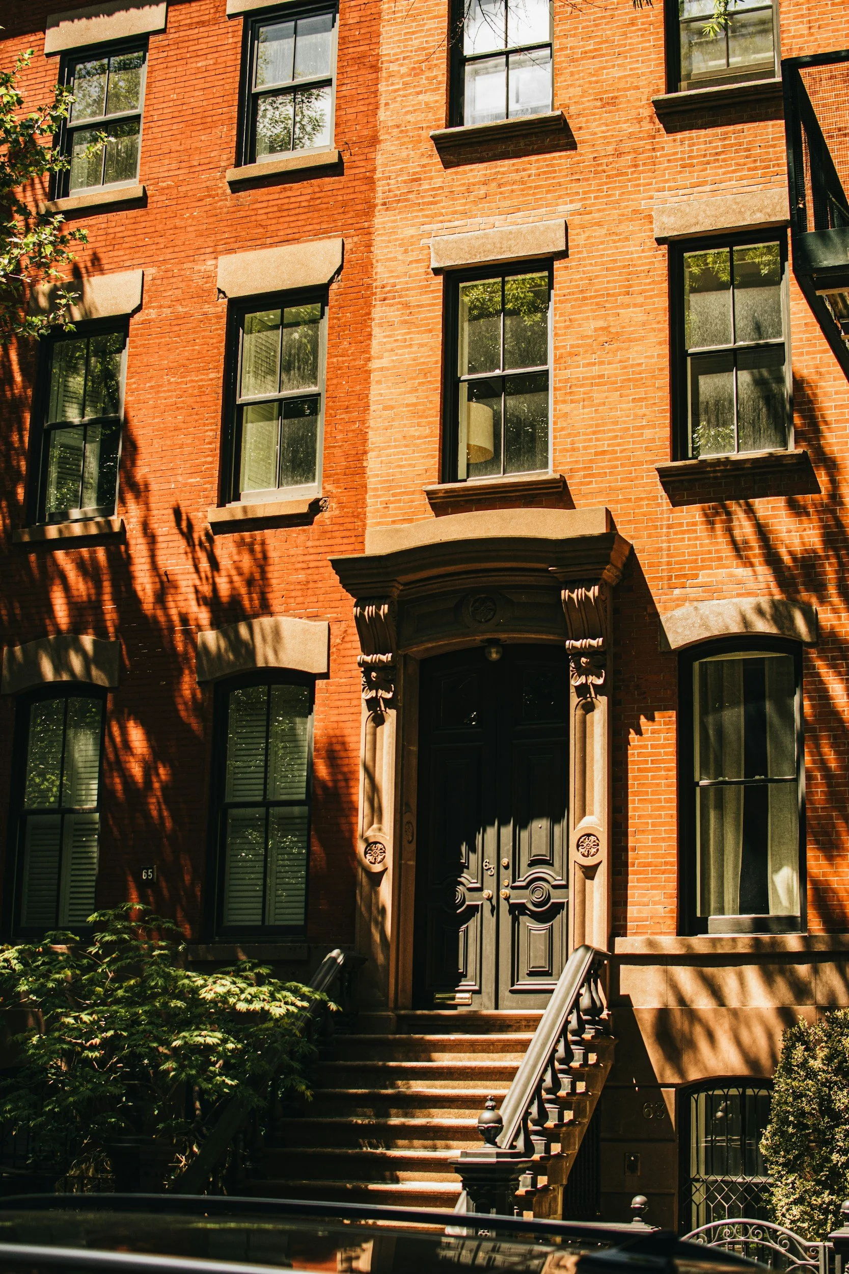 A multi-story brick residential building with tall windows, stone window sills, and an ornate doorway with stairs and black railings. Shadows of nearby trees can be seen on the facade.