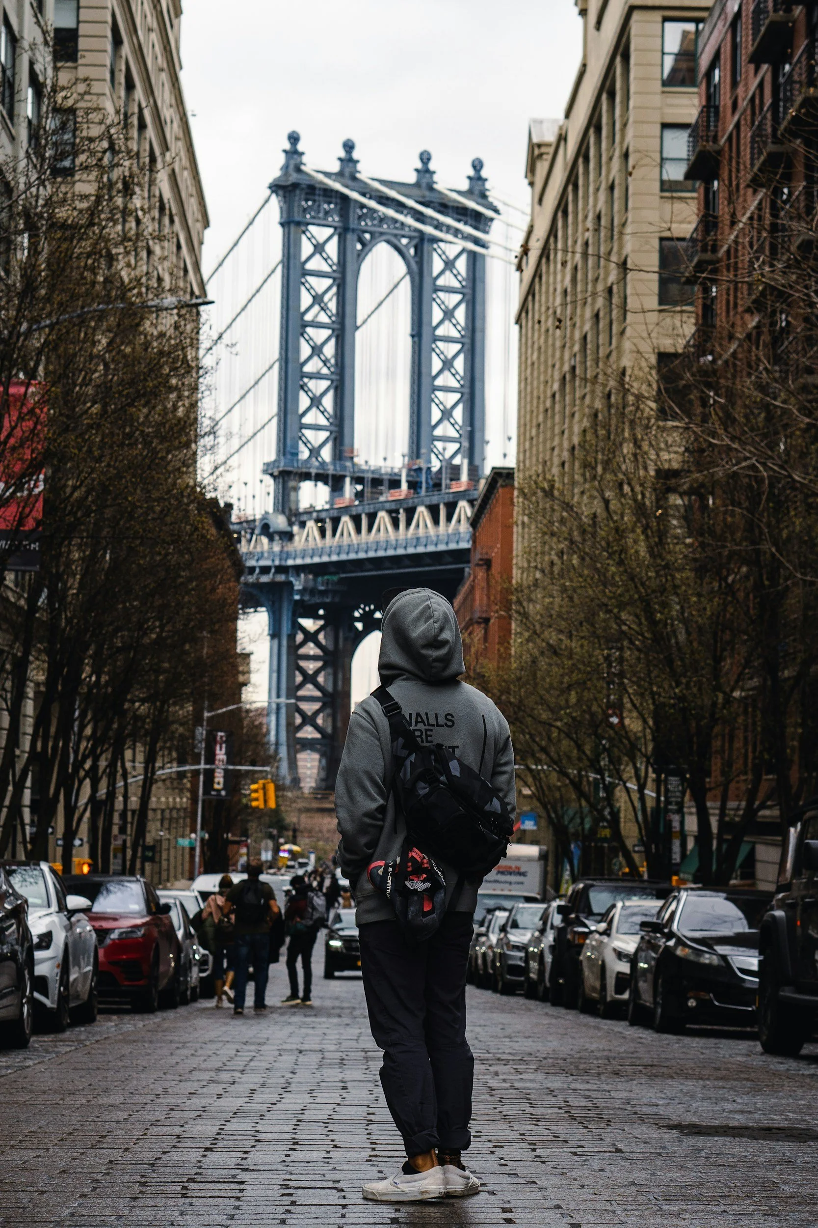 Person wearing a gray hoodie with a backpack, walking on a cobblestone street, with the Brooklyn Bridge in the background, surrounded by parked cars and other pedestrians.