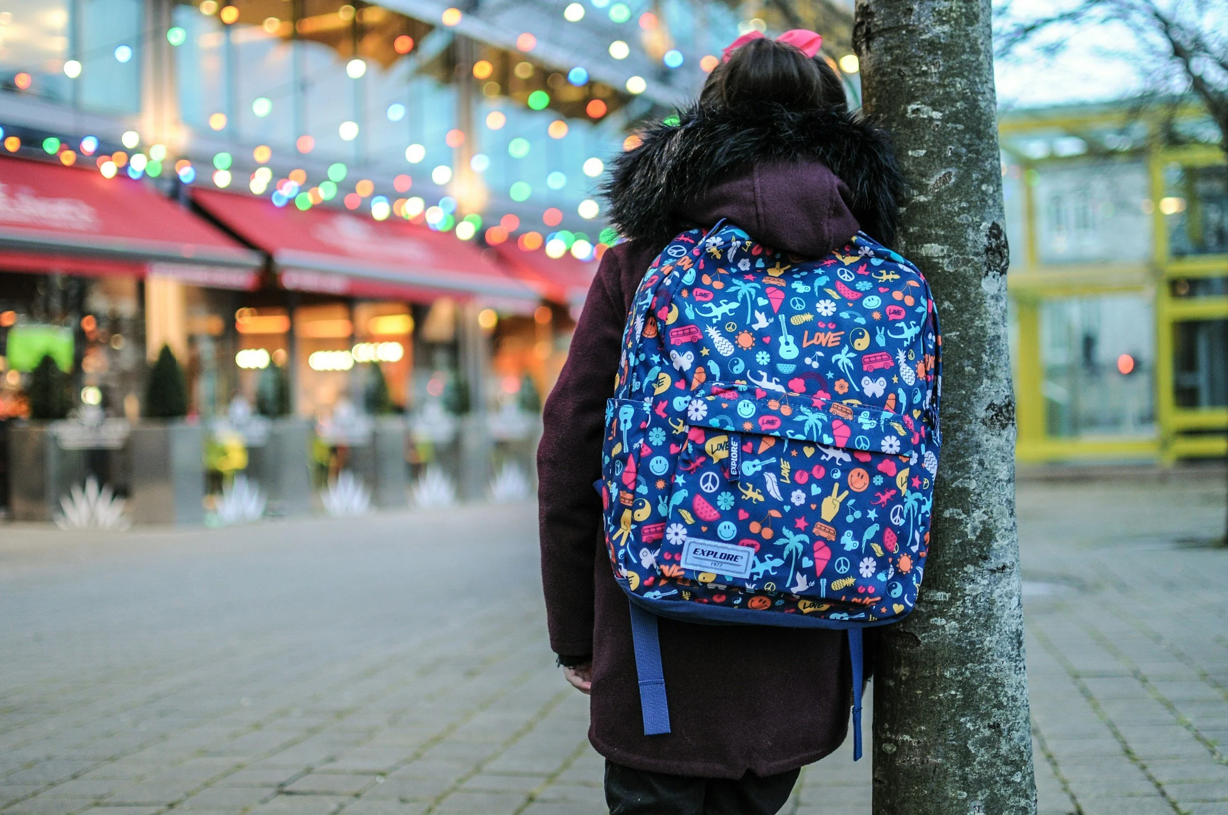 A child with a pink bow in her hair, wearing a dark coat with a faux fur collar, stands with her back to the camera behind a tree. She has a colorful backpack with doodle-like designs, including smiley faces, peace signs, and the word 'LOVE'. The scene is outdoors with string lights and a blurred background of shops or stalls.