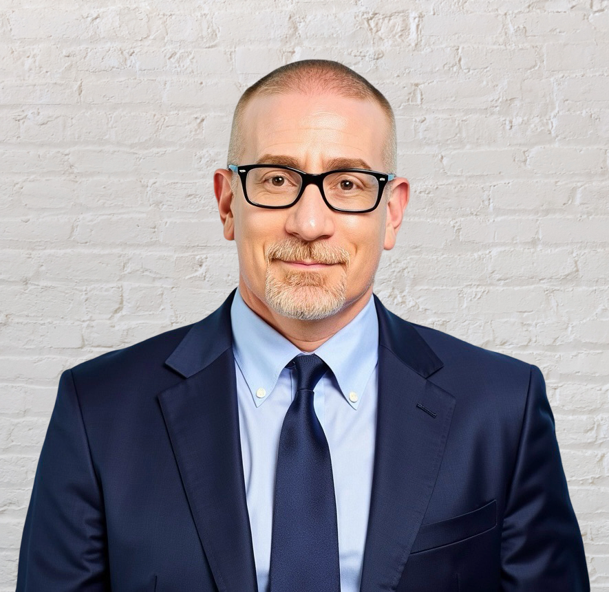 A man with short hair, glasses, and a beard wearing a navy suit, light blue shirt, and navy tie standing against a white brick wall.