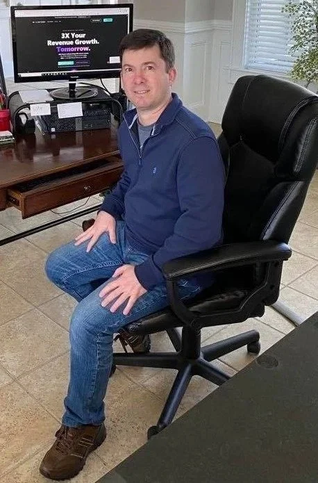 A man sitting in a black office chair in a home office, with a desk, computer, and window with blinds in the background.