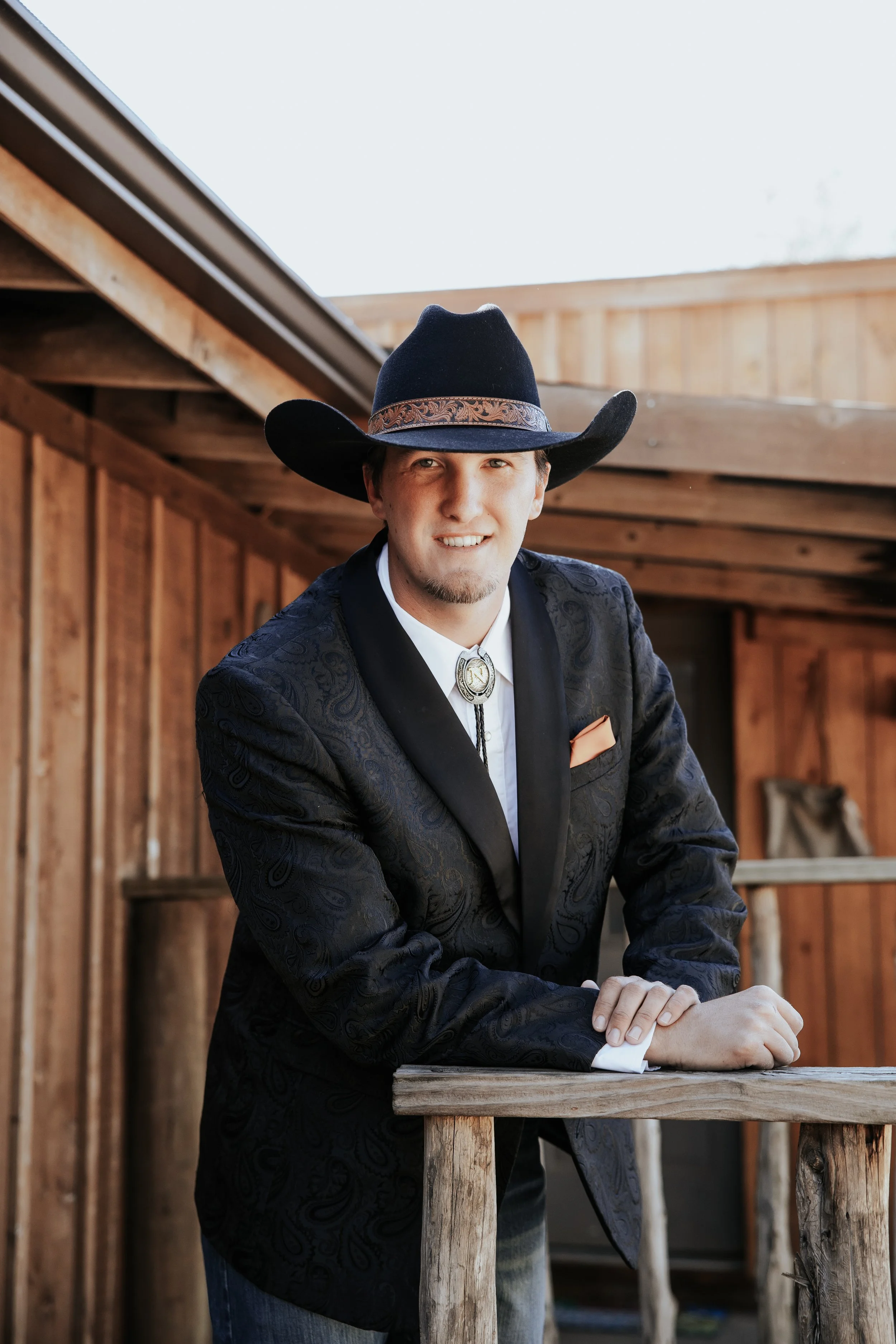 A man in a cowboy hat and western attire leaning on a wooden railing in a rustic barn setting.