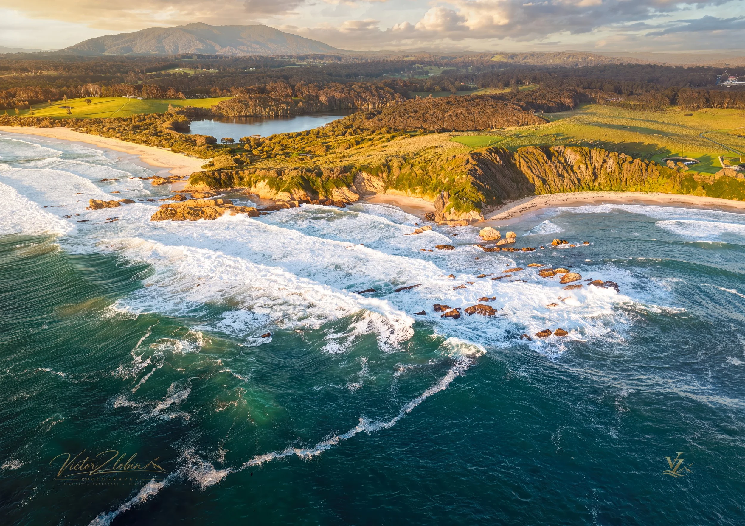 Aerial coastal landscape of Australia at golden hour, featuring rugged cliffs, sandy beaches, turquoise ocean, green countryside and distant mountains