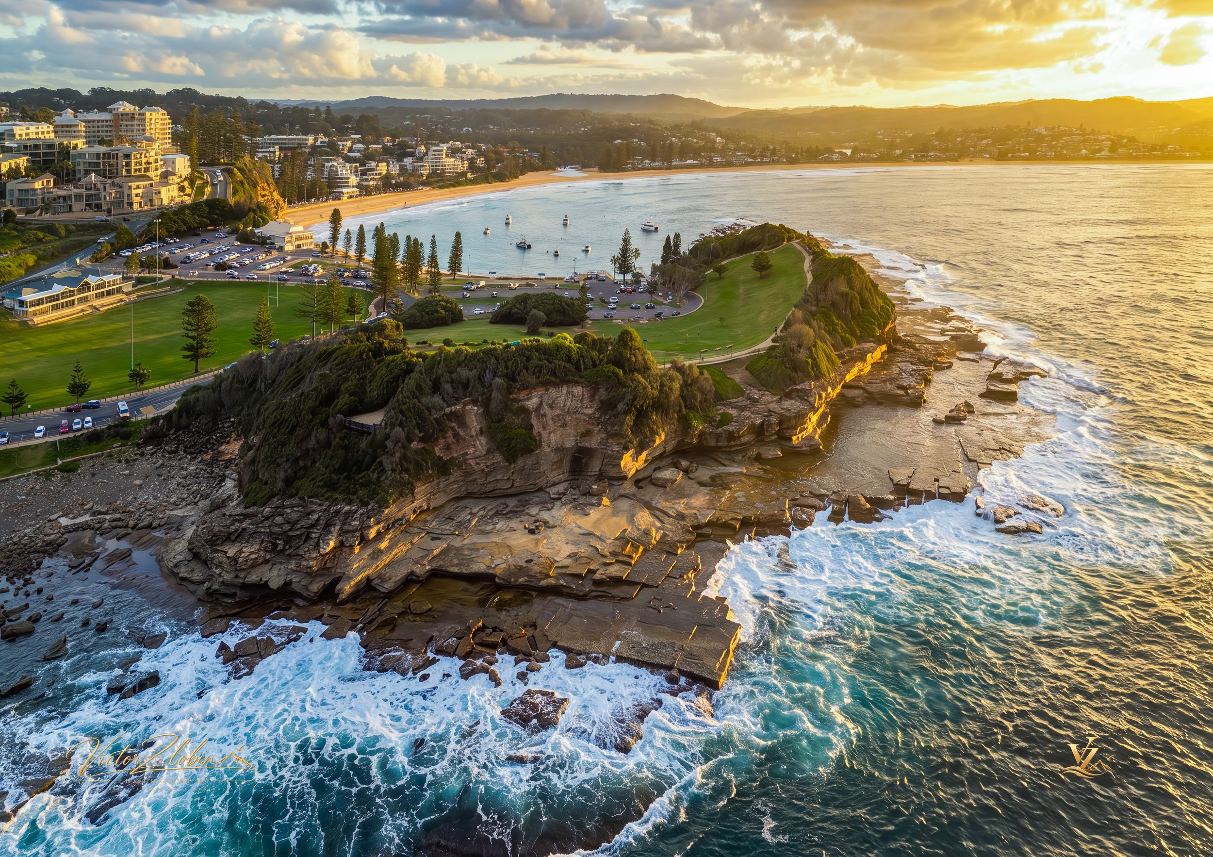 Aerial photograph of Terrigal Headland on the Central Coast of NSW, showcasing dramatic sandstone cliffs, rugged ocean rock platforms and turquoise waves glowing in warm golden-hour light. 0674