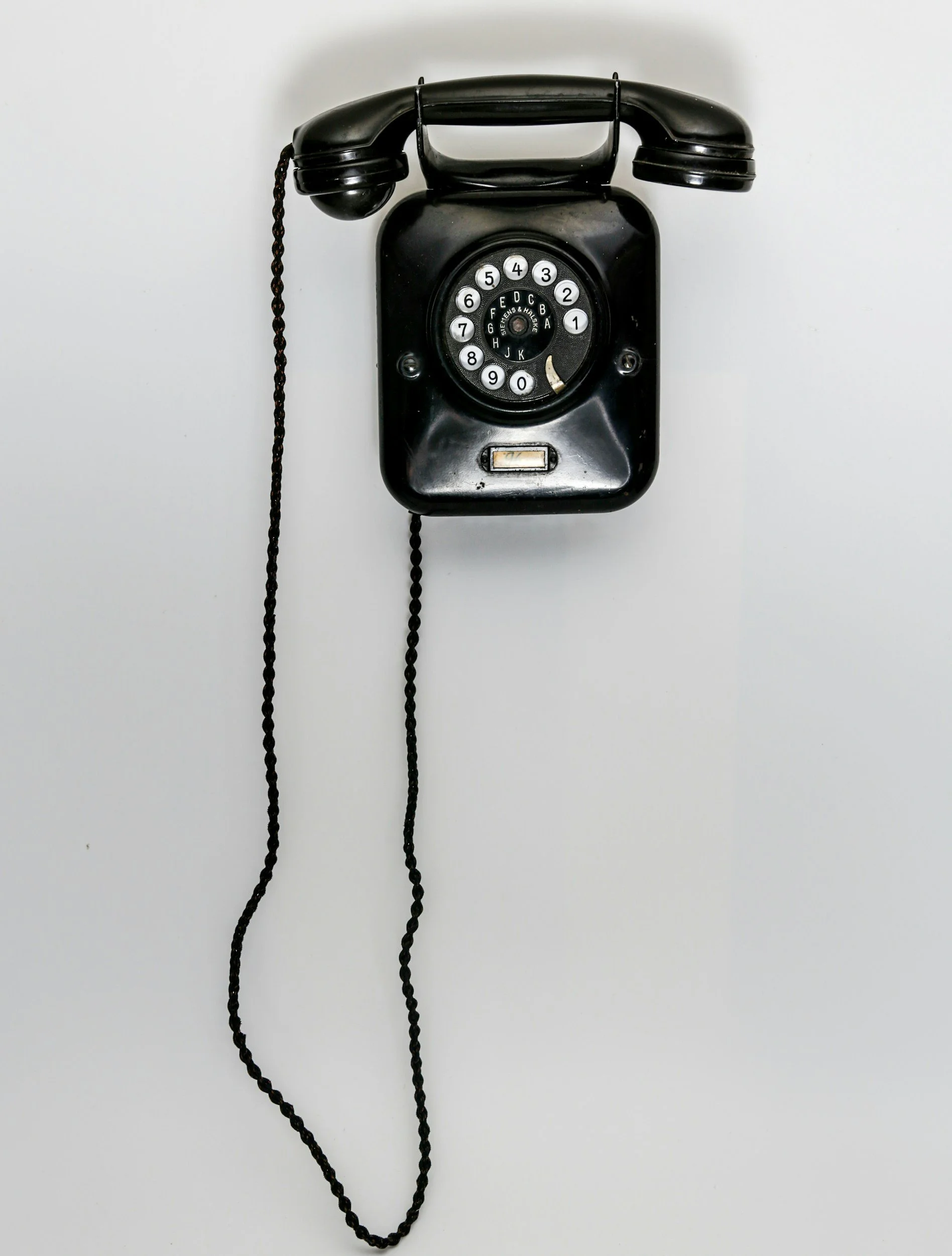 Black rotary dial telephone mounted on a white wall, with black coiled cord hanging down.