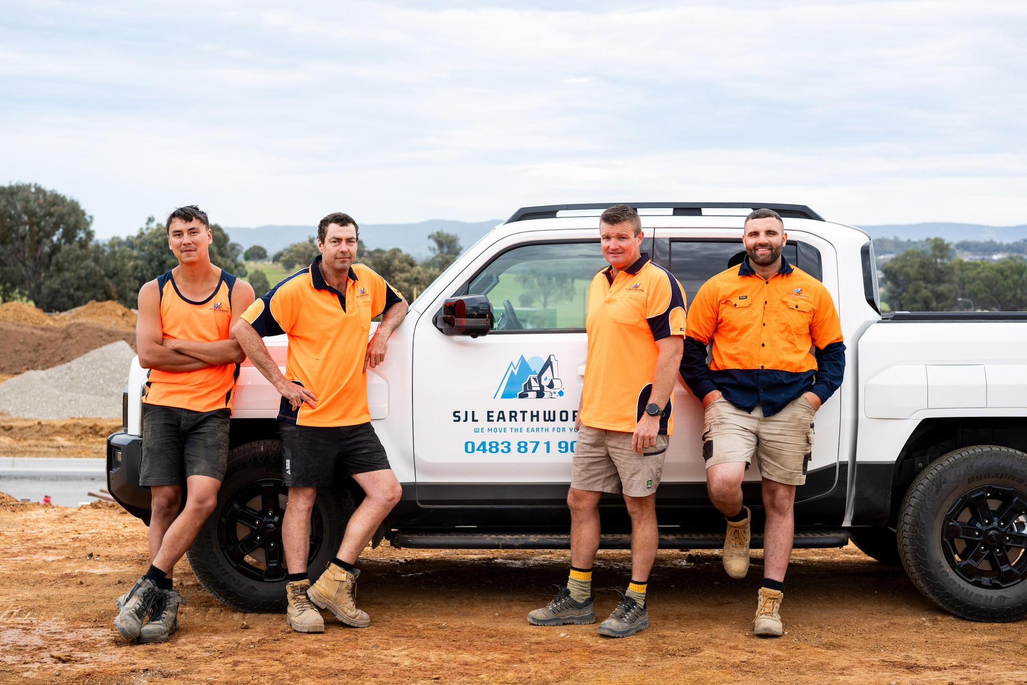 Four construction workers standing next to a white truck with company logo at construction site, with soil and trees in background.
