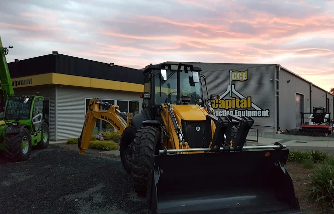 A yellow New Holland backhoe loader is parked outside a building with a sign that reads 'Capital Equipment.' The sky is pink and purple with clouds.