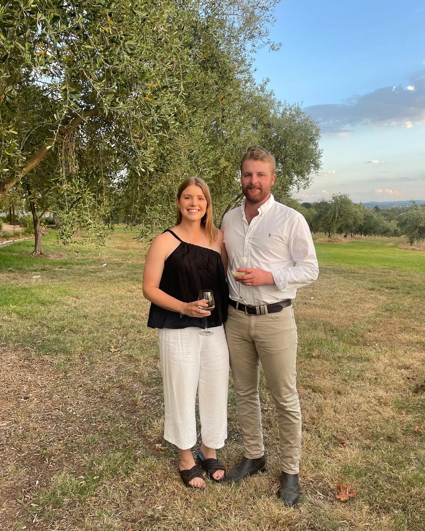 A young woman and a young man standing outdoors in a grassy area with trees and a blue sky in the background. They are holding glasses of wine and smiling at the camera, dressed casually.