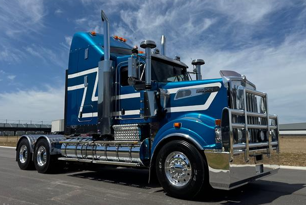 Blue semi-truck with chrome accents parked on a road under a partly cloudy sky.