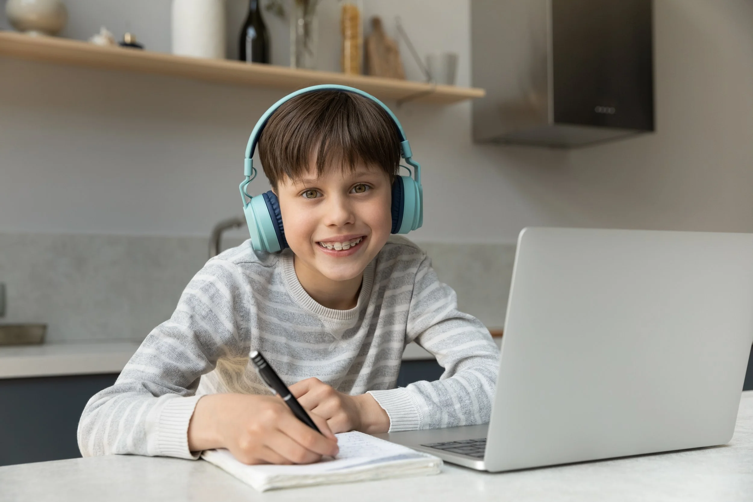 Young boy with brown hair and green eyes, wearing a gray striped sweater and blue headphones, smiling while writing in a notebook, sitting at a kitchen counter with a laptop in front of him.