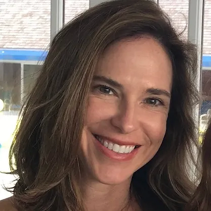 Smiling woman with brown hair and a bright expression, in an indoor setting with windows in the background.